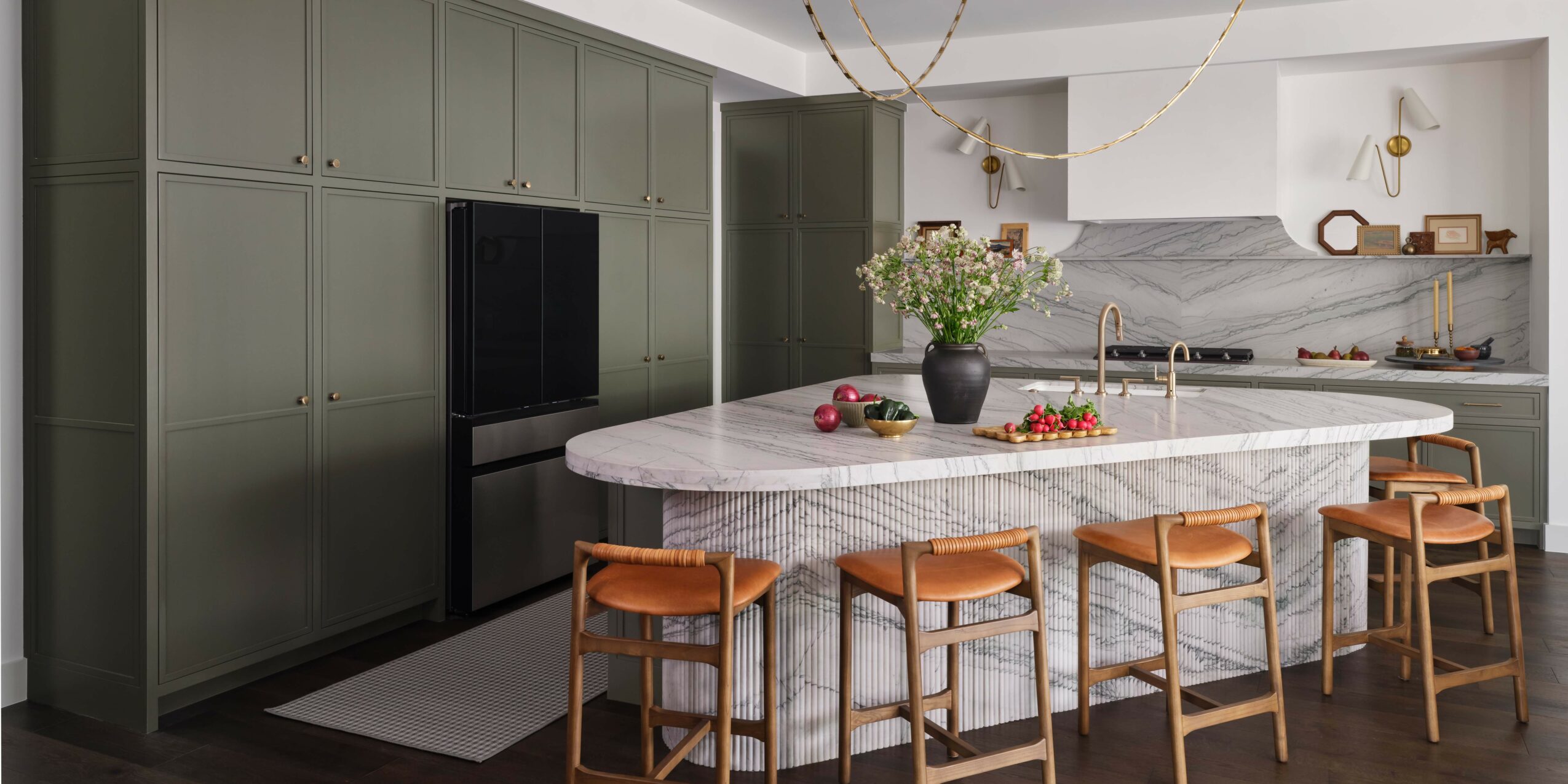 Modern kitchen with green cabinets, marble island, and bar stools, decorated with flowers and hanging lights.