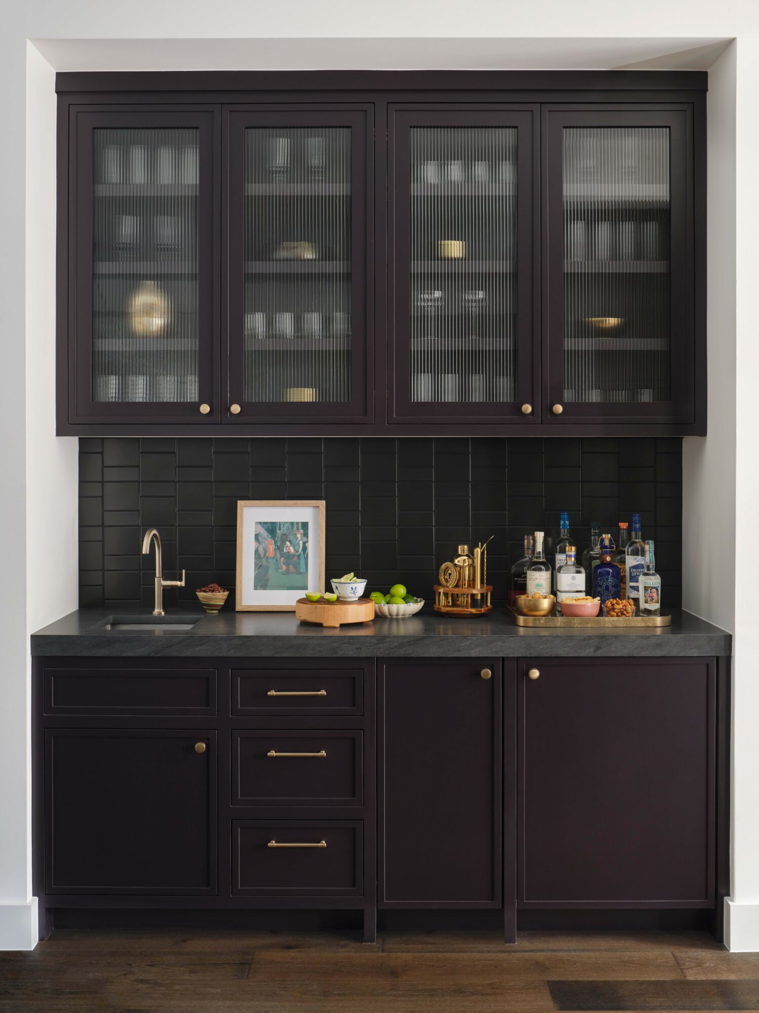 Kitchen cabinet with glass doors, black countertop, sink, and various bottles and items on the counter, dark wood flooring.