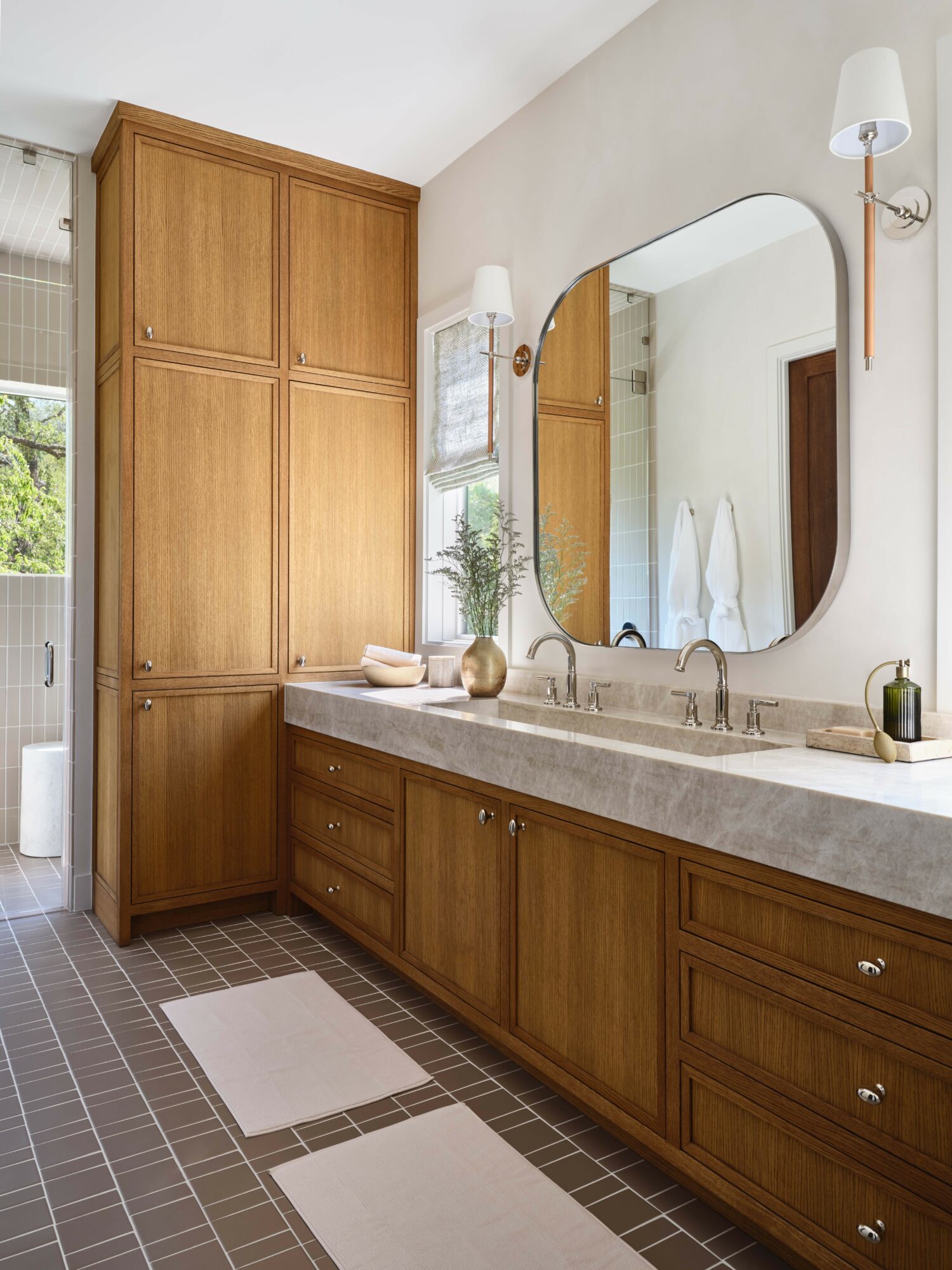 Bathroom vanity with wooden cabinets, large mirror, and a beige countertop, with a window and a wooden door in the background.