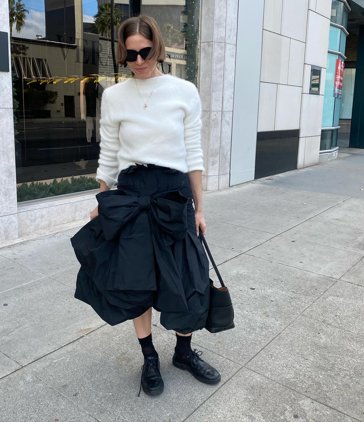 Woman in white sweater, black bag, sunglasses, and black boots standing on sidewalk outside building.