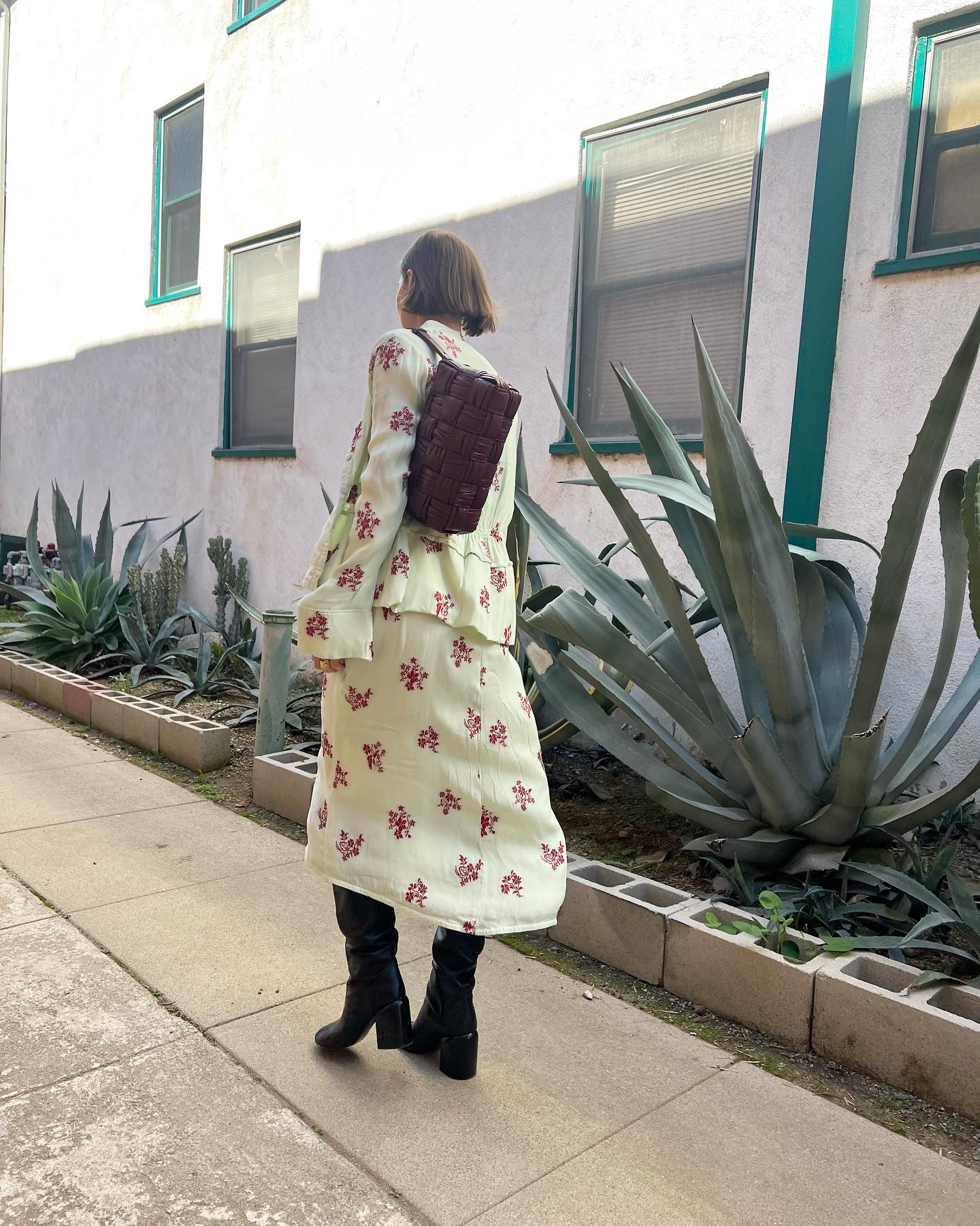 Person walking along sidewalk with large agave plants and a white building in background.
