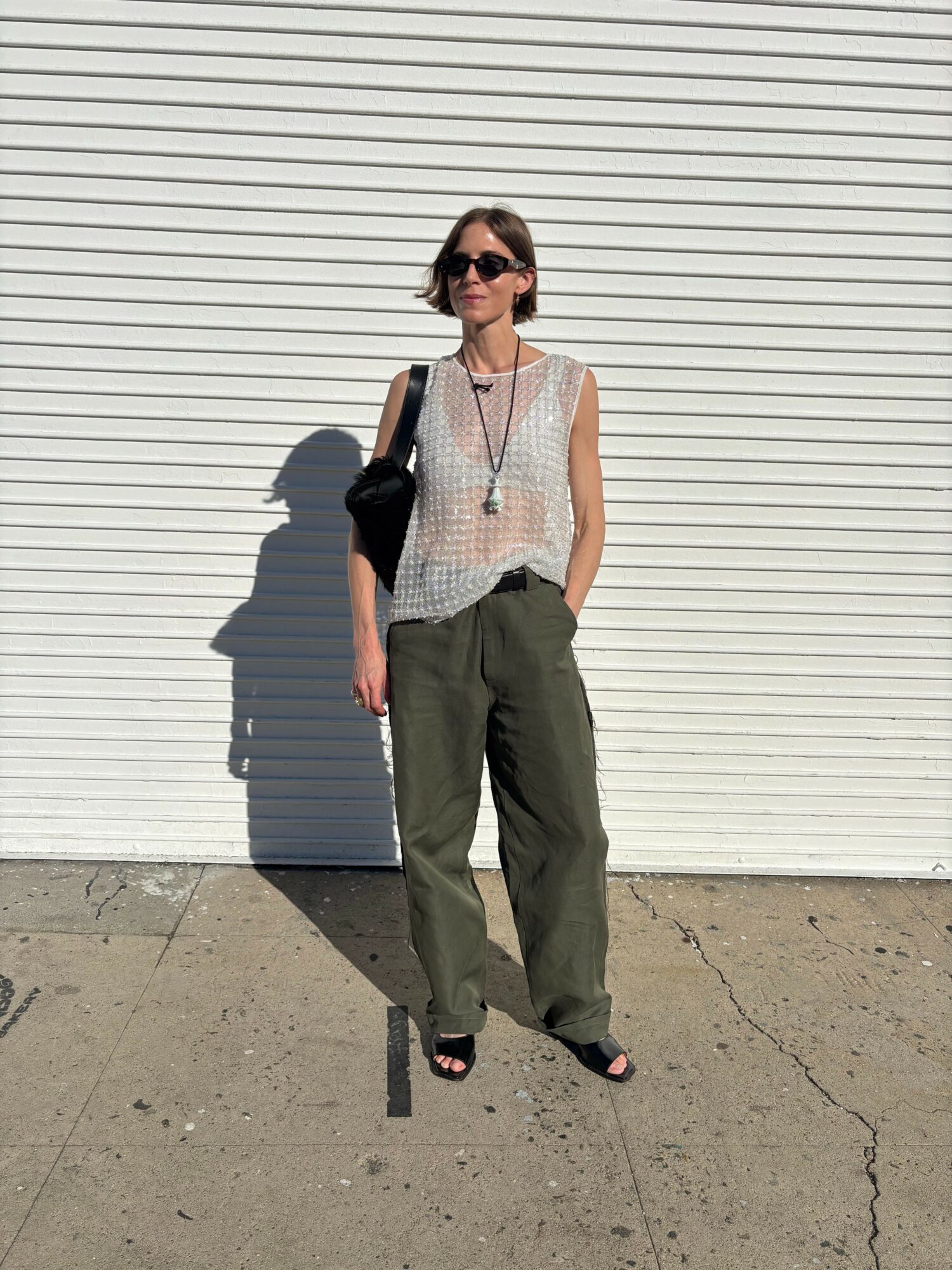Woman standing outdoors against a white corrugated wall, wearing sunglasses, a sleeveless top, and wide-leg pants, holding a bag.