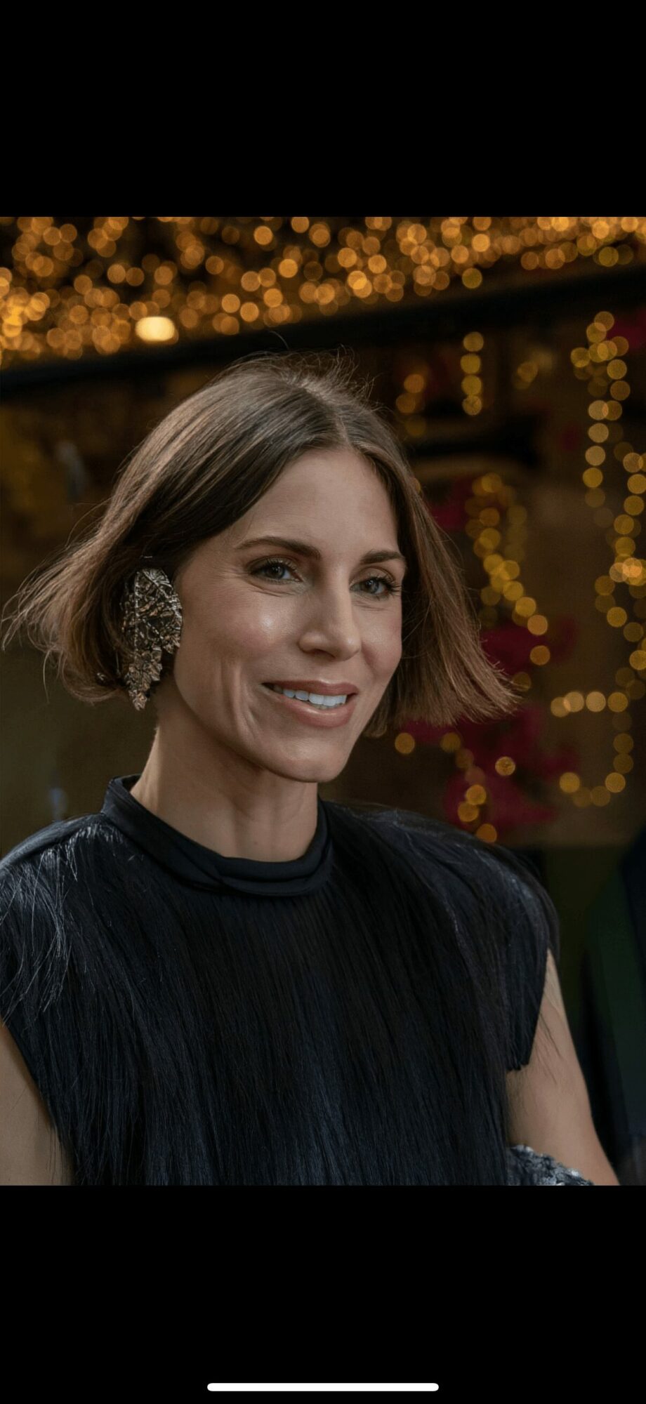 Woman with short hair and earrings smiling at an event with blurred golden lights in background.