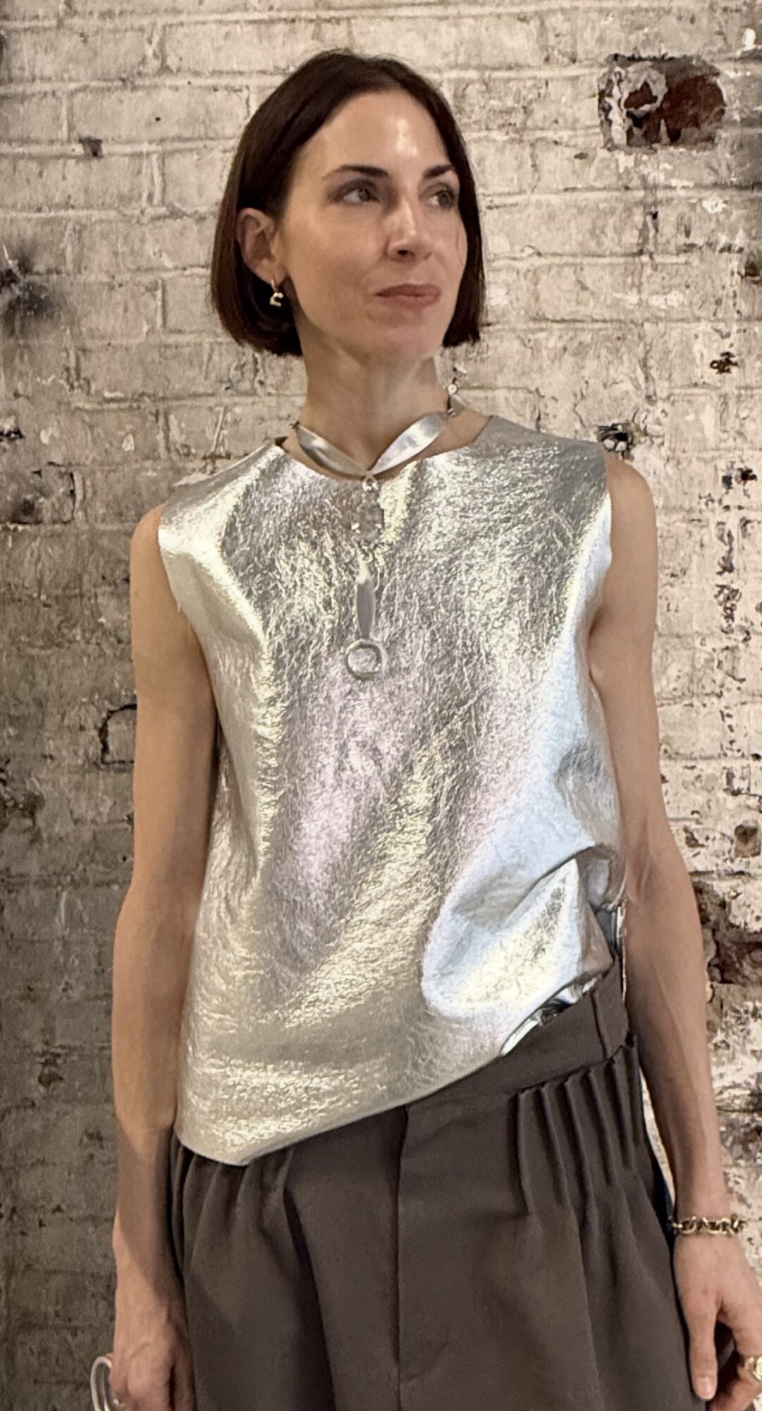 Woman with short dark hair wearing a shiny silver sleeveless top and dark pants, standing against a brick wall.