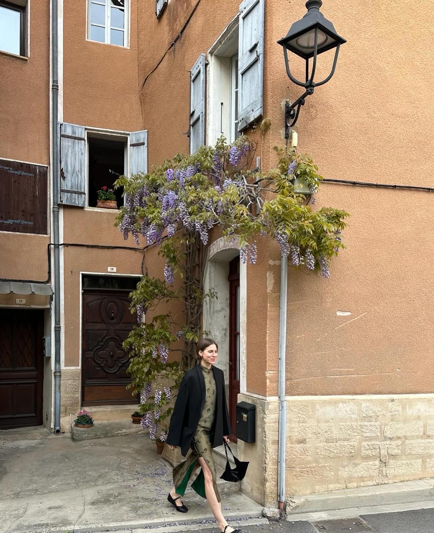 Woman walking on a street with a building, purple flowering plant, and street lamp in the background.