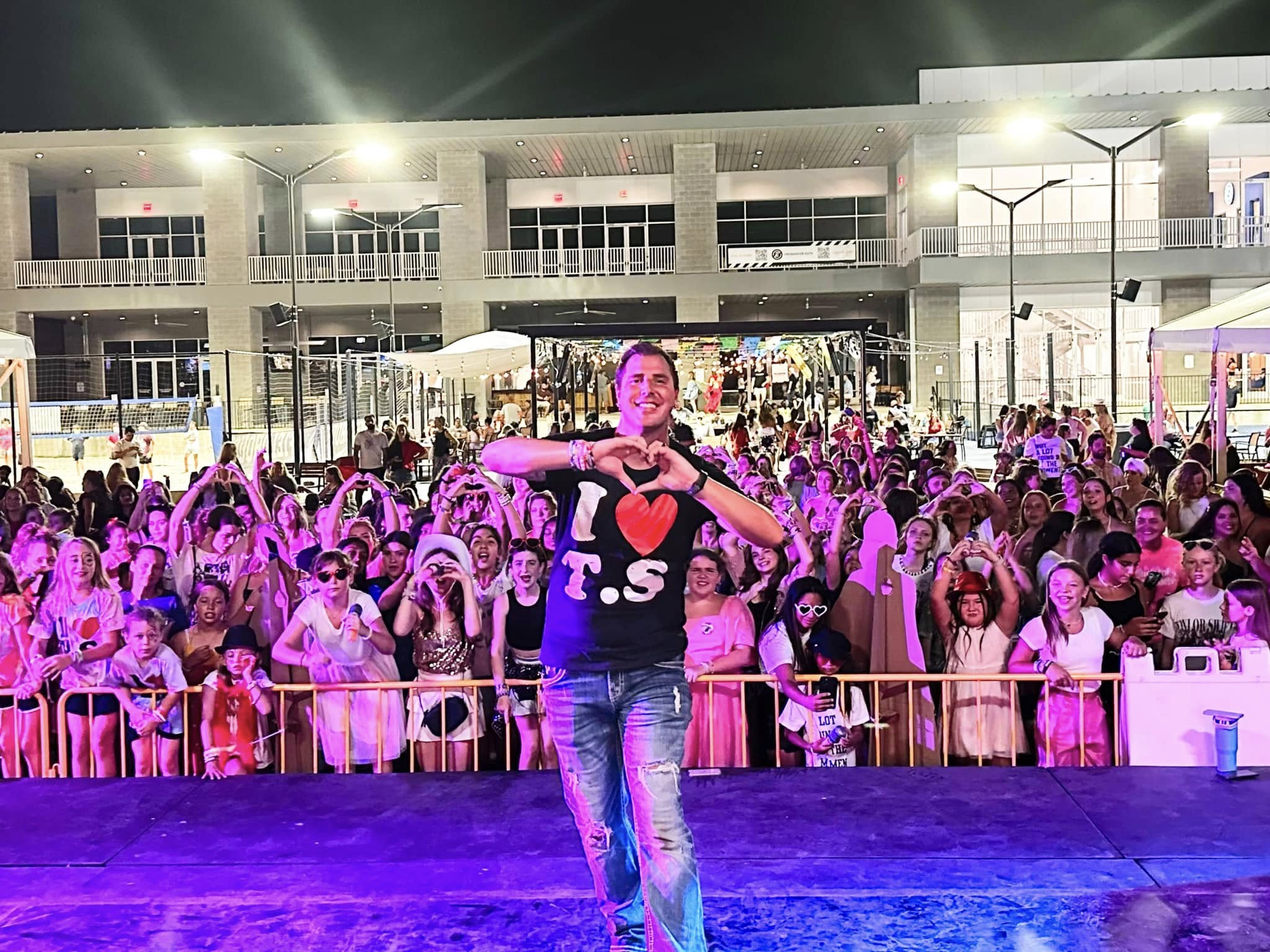 Person on stage with a large crowd behind, building with balconies in the background, night scene, colorful lighting.