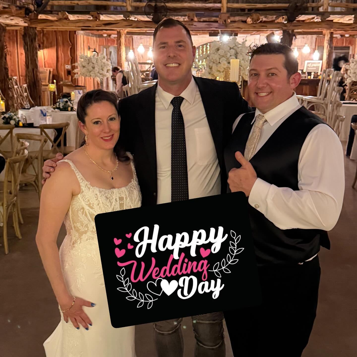 Three people smiling at a wedding reception, holding a sign that says 'Happy Wedding Day,' in a decorated venue.