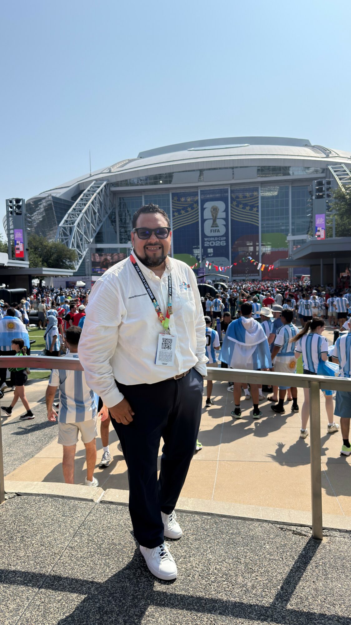 Man wearing sunglasses, white shirt, and dark pants standing outdoors in front of a large stadium with a crowd.