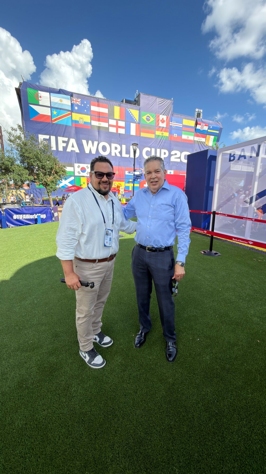 Two men standing on grass in front of FIFA World Cup signage, smiling, with blue sky and clouds above.