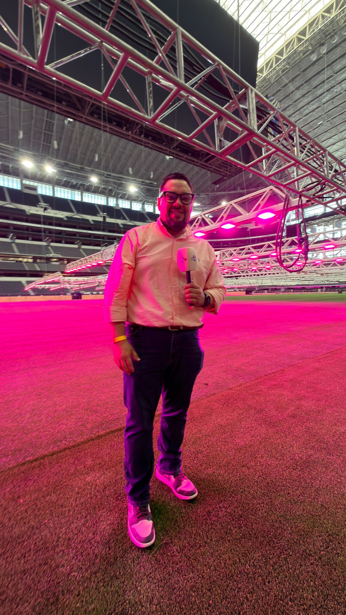 Man standing on a field inside a large stadium, holding a microphone, with pink lighting and structural beams overhead.
