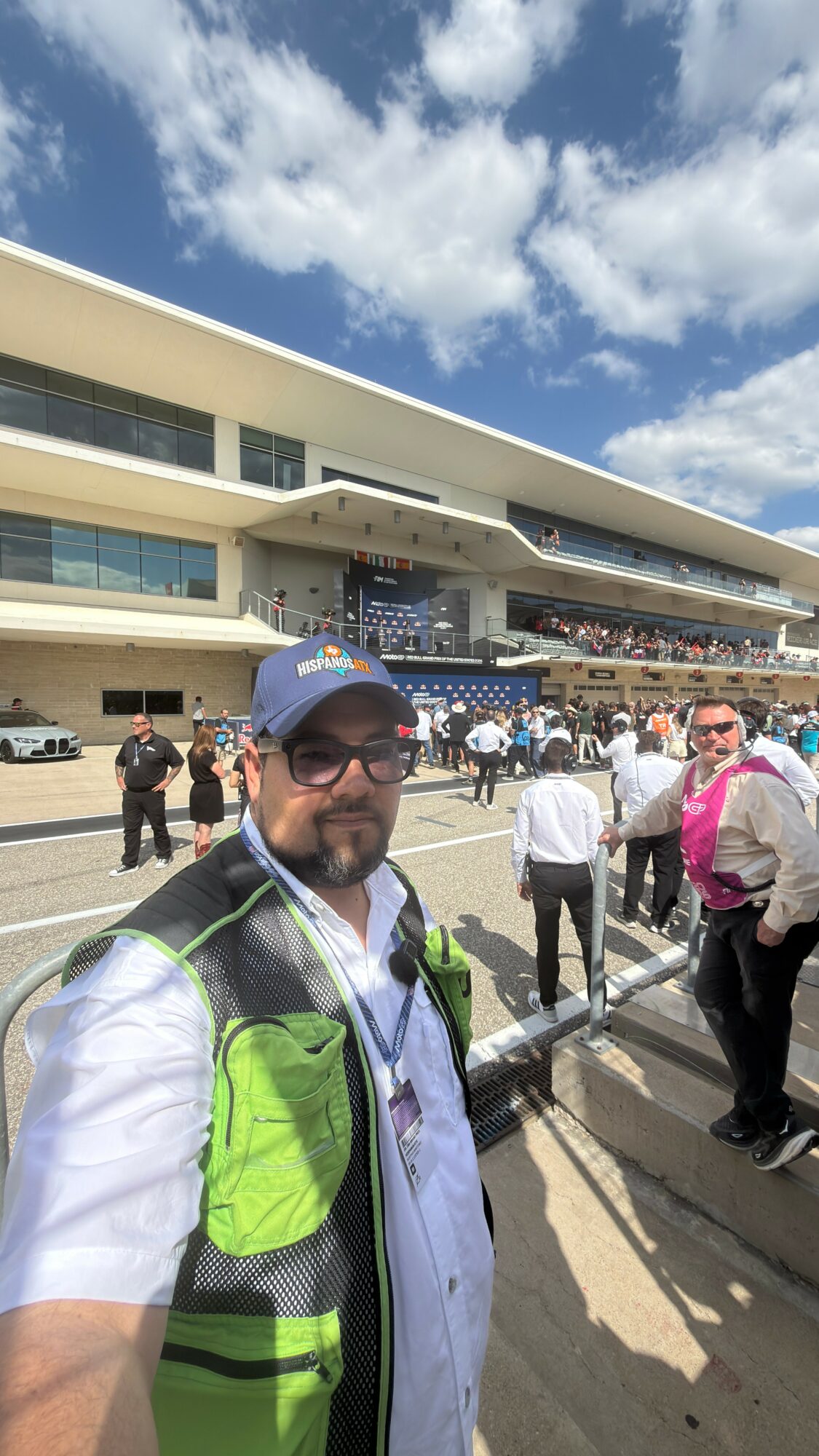 Man wearing sunglasses, a blue cap, and a backpack taking a selfie at a busy outdoor event with a large building and crowd in background.