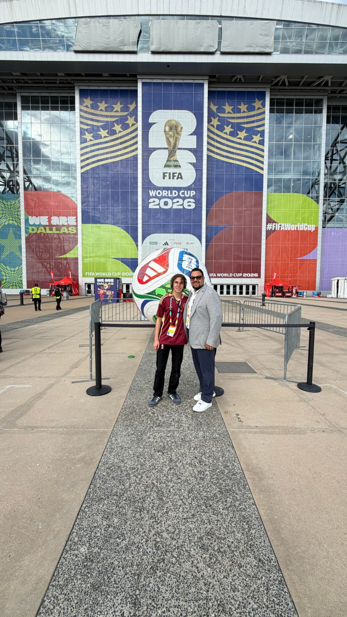Two people standing in front of a large FIFA World Cup 2026 banner outside a building, with a gray walkway and black stanchions.