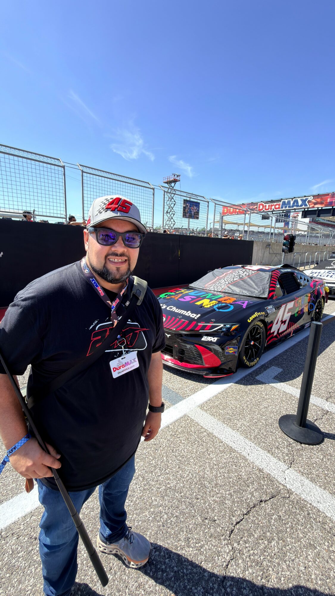 Man with sunglasses and a cap standing next to a race car on a track, with a clear blue sky above.