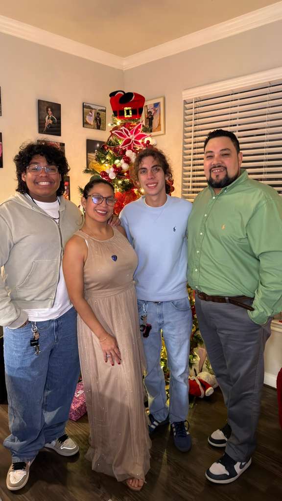 Four people standing in front of a decorated Christmas tree, smiling indoors, with a window and wall art in the background.