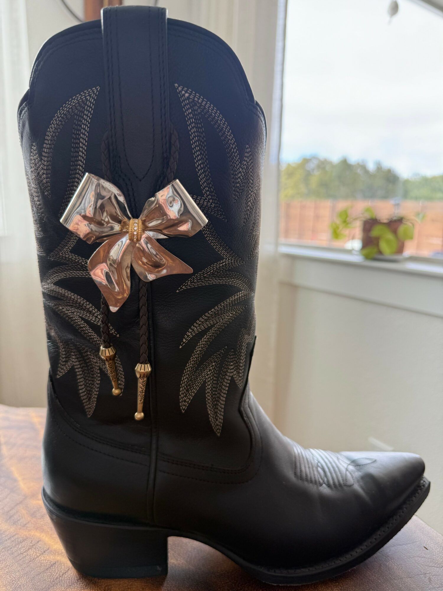 Black cowboy boot with a gold bow decoration, placed on a wooden surface near a window with a view outside.
