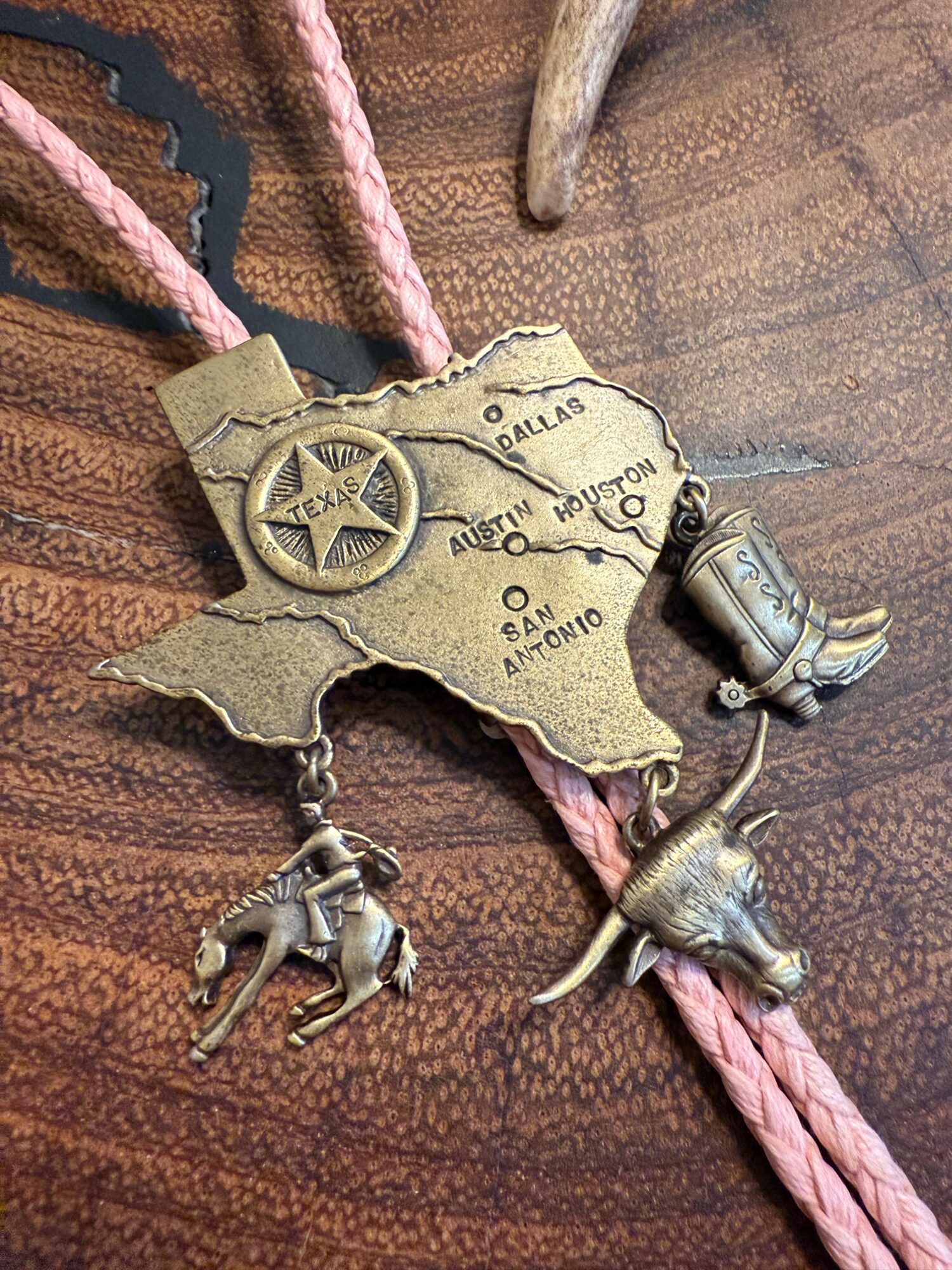 Metal Texas-shaped pendant with a star, map details, and hanging cowboy boot and horse charms, attached to pink braided cord.