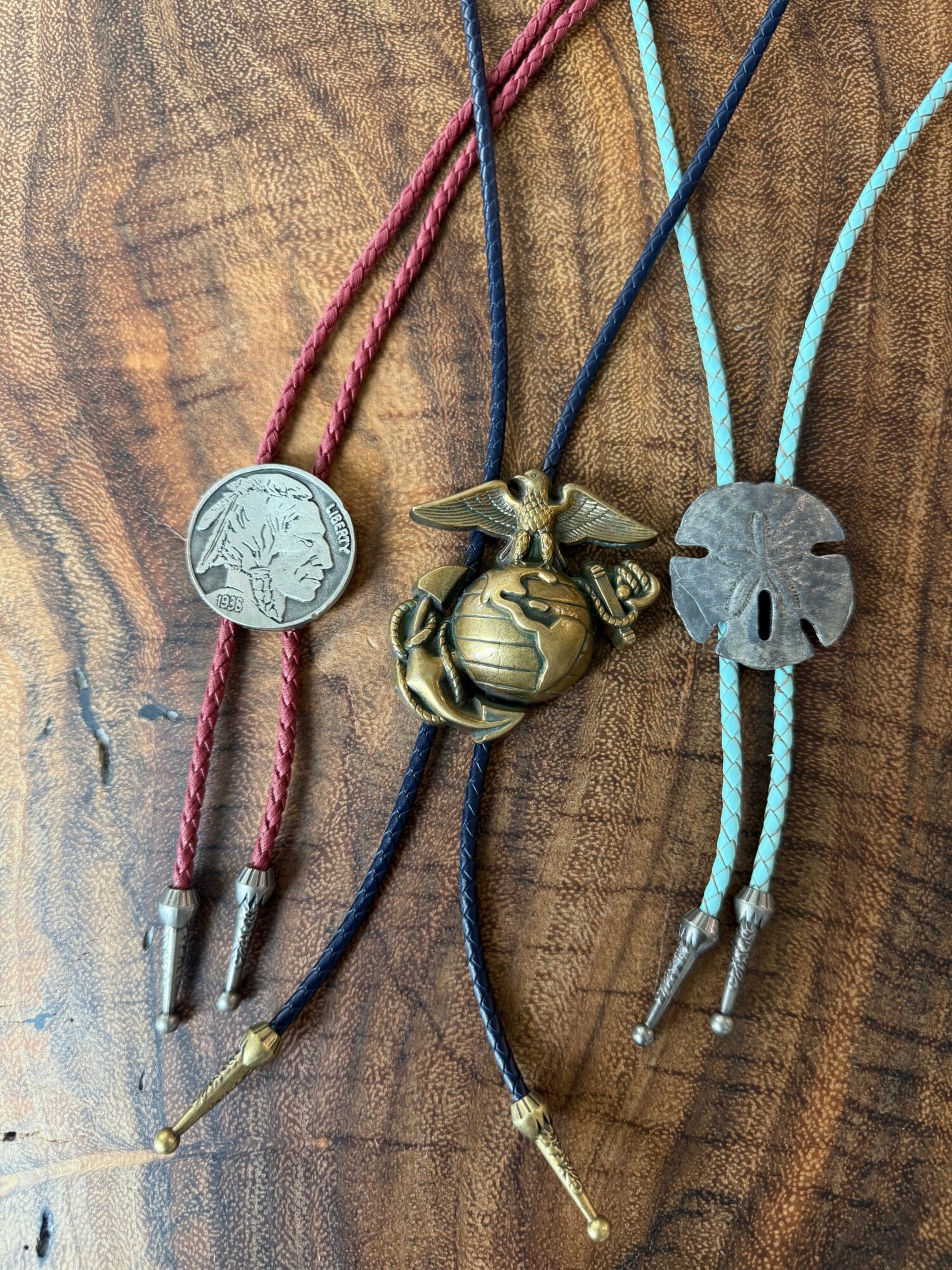 Four necklaces with pendants and cords on a wooden surface, featuring a coin, a brass eagle, and a round pendant.