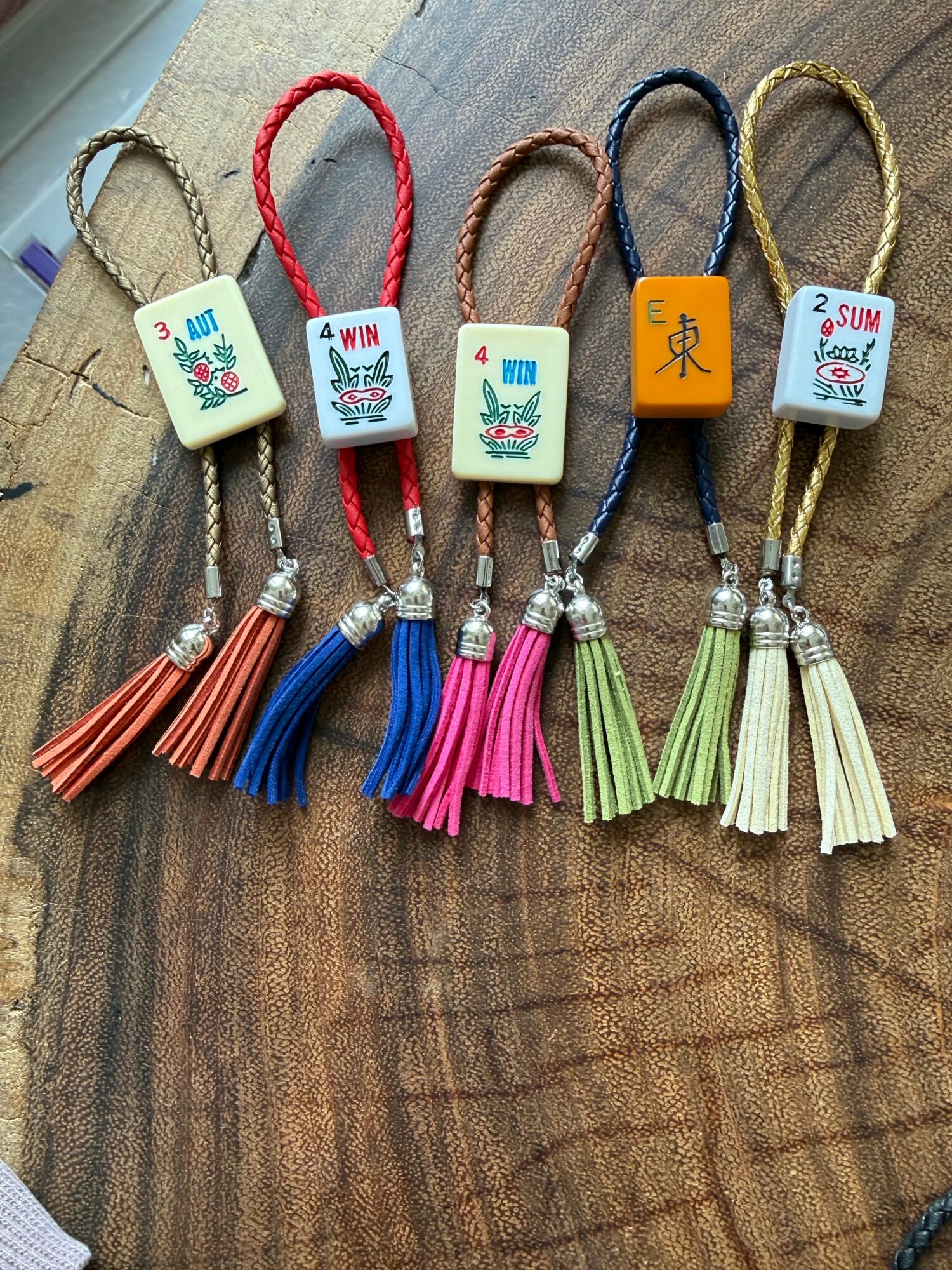 Five decorative cords with tags and tassels, arranged on a wooden surface, each with different colors and labels.