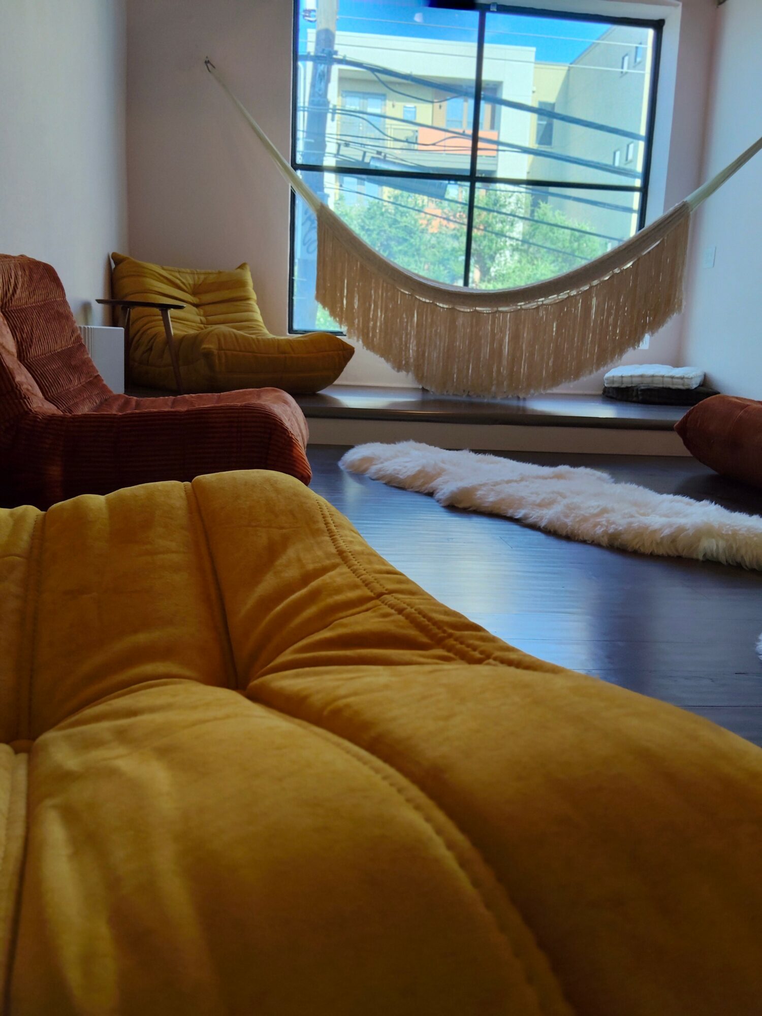 Living room with colorful chairs, a hammock by the window, and a rug on dark wooden floor.