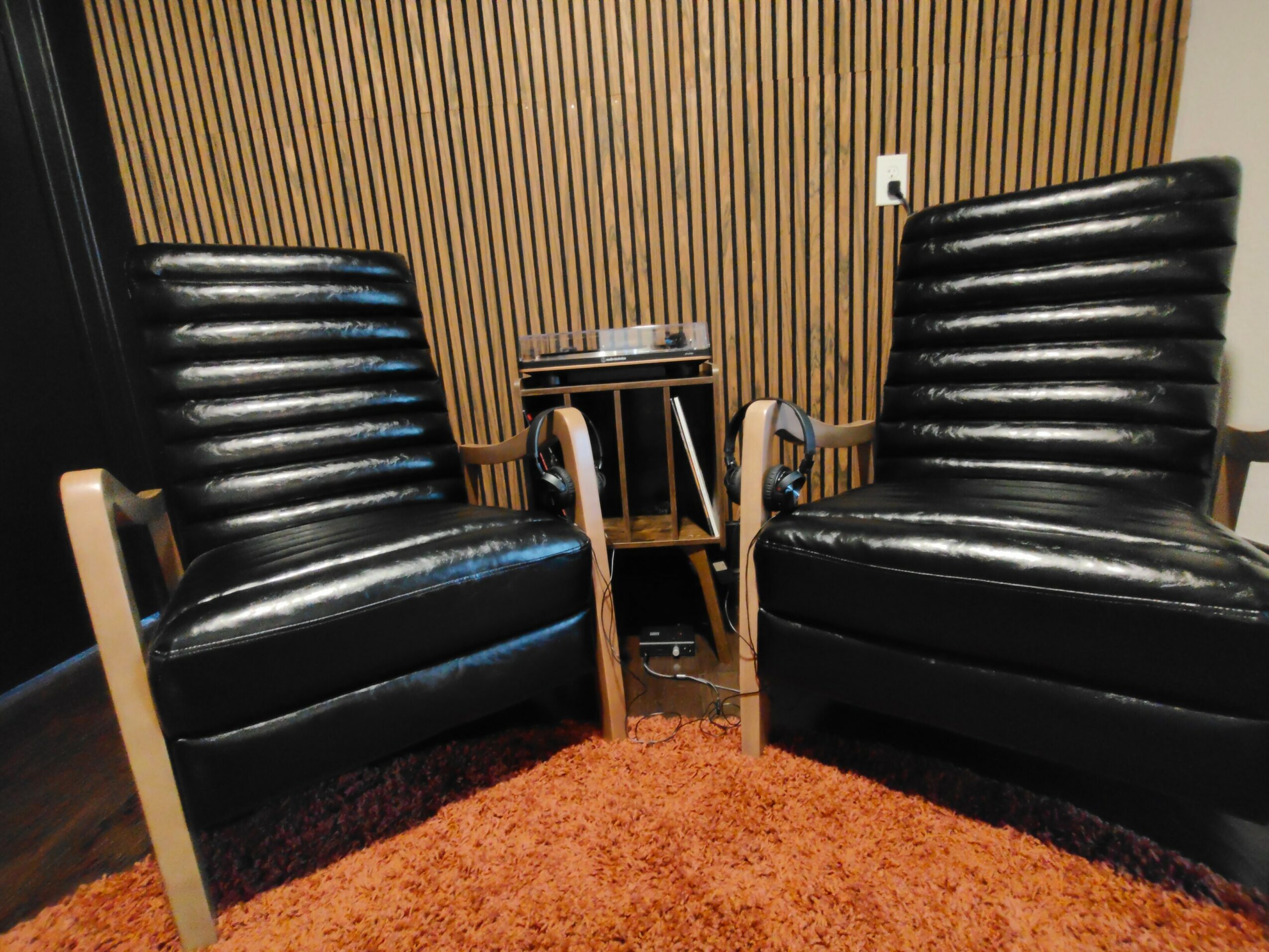 Two black leather armchairs with wooden armrests and a small table between them, in a room with wooden wall paneling and a reddish-brown carpet.