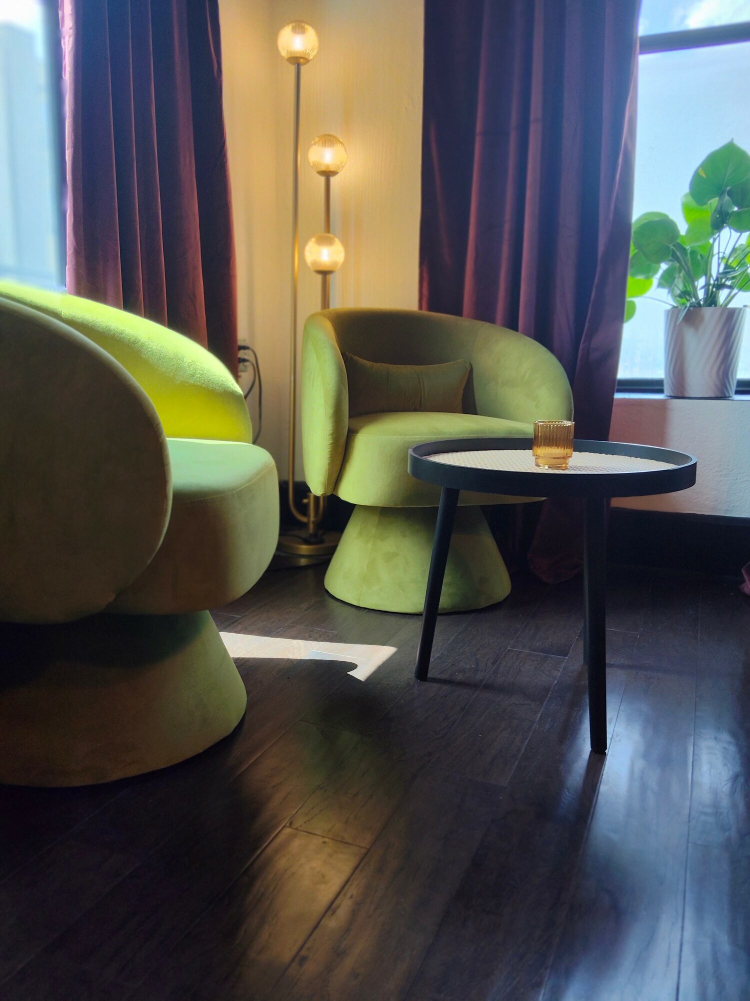Interior scene with two green chairs, a small black table, a window with potted plants, and a floor lamp, in a cozy room.