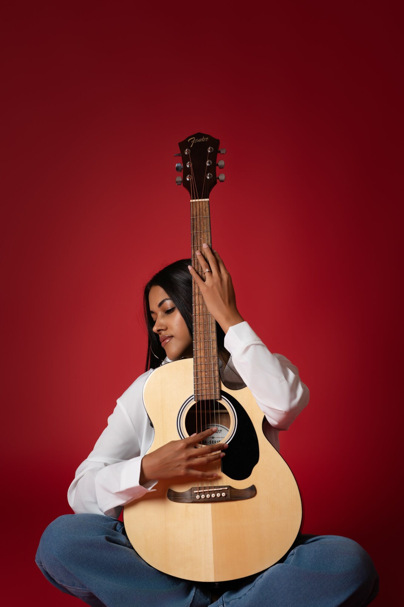Person sitting cross-legged holding an acoustic guitar against a red background.