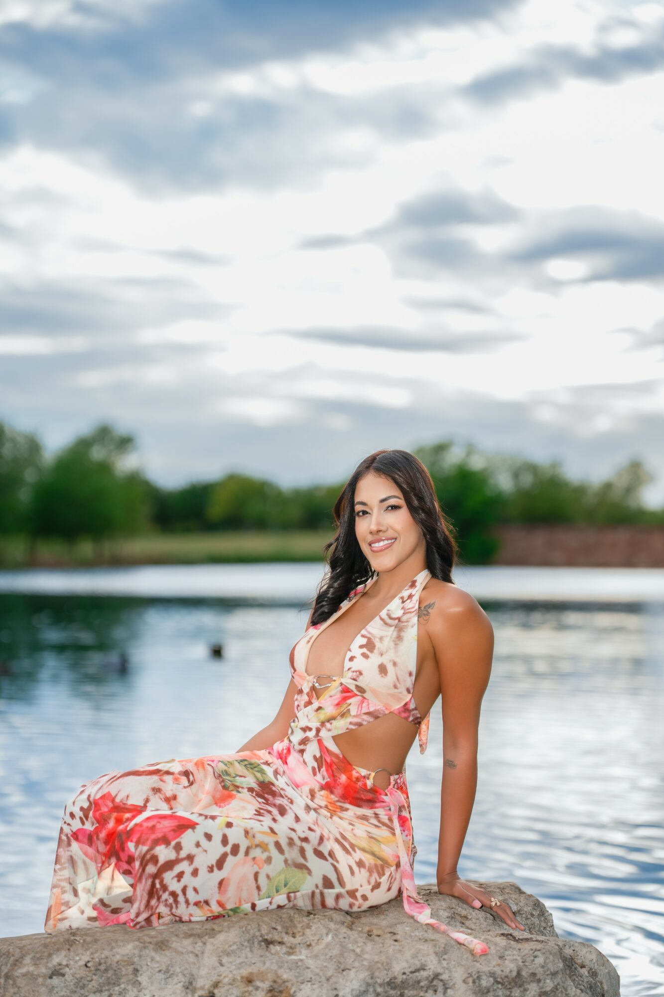Woman sitting on rocks by water, smiling, wearing a floral dress, outdoors under cloudy sky.