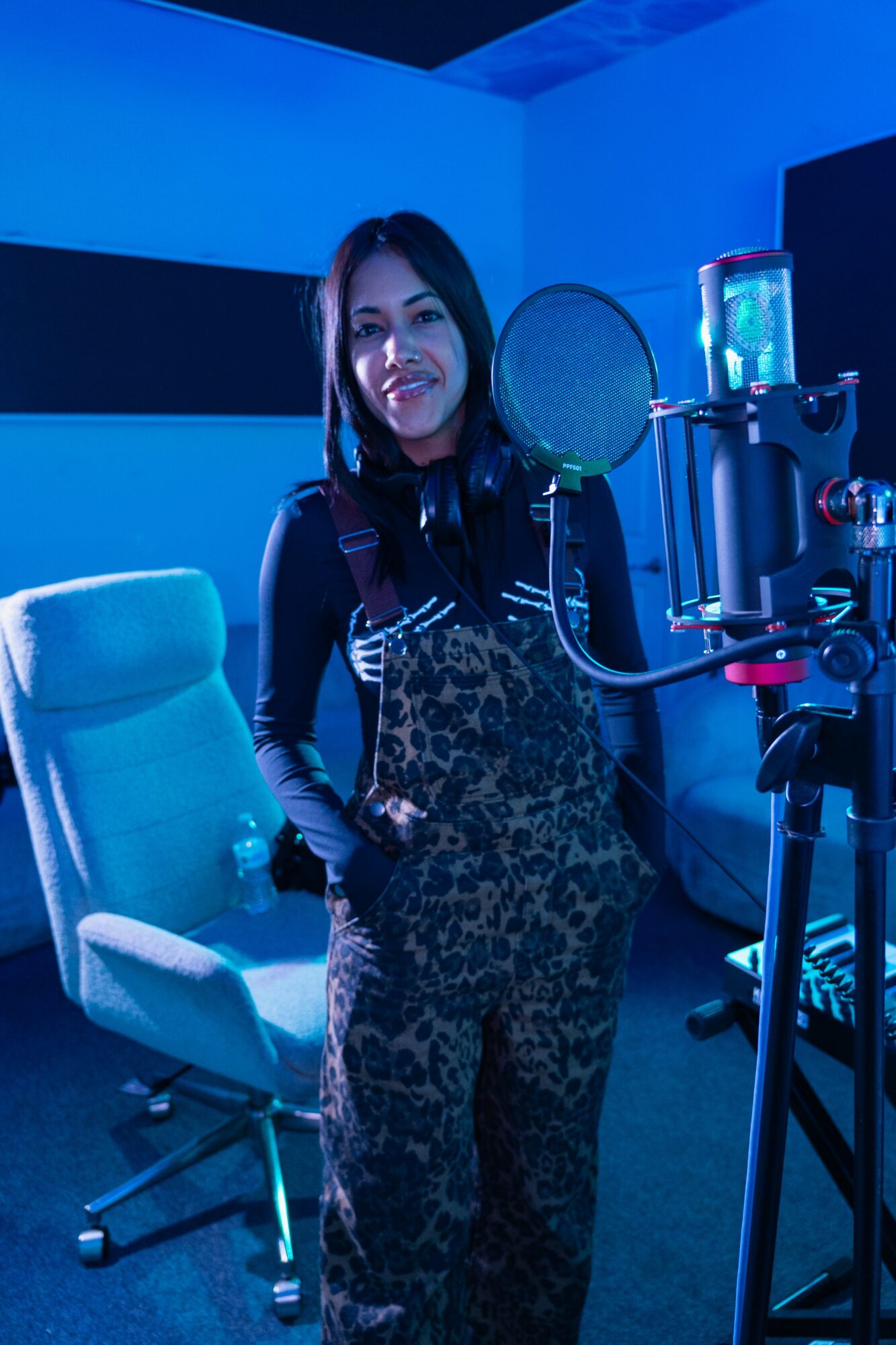 Woman in a recording studio with microphone, chair, and equipment, smiling at the camera.