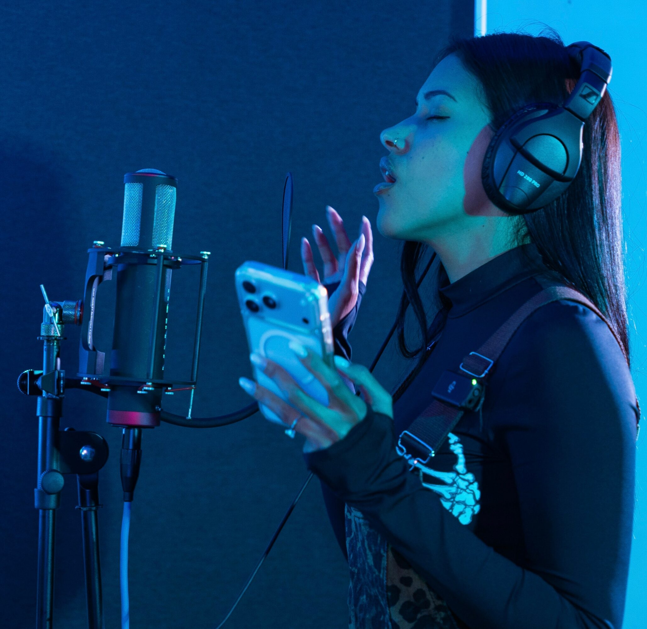 Woman in headphones singing into a microphone while holding a smartphone, with blue lighting.