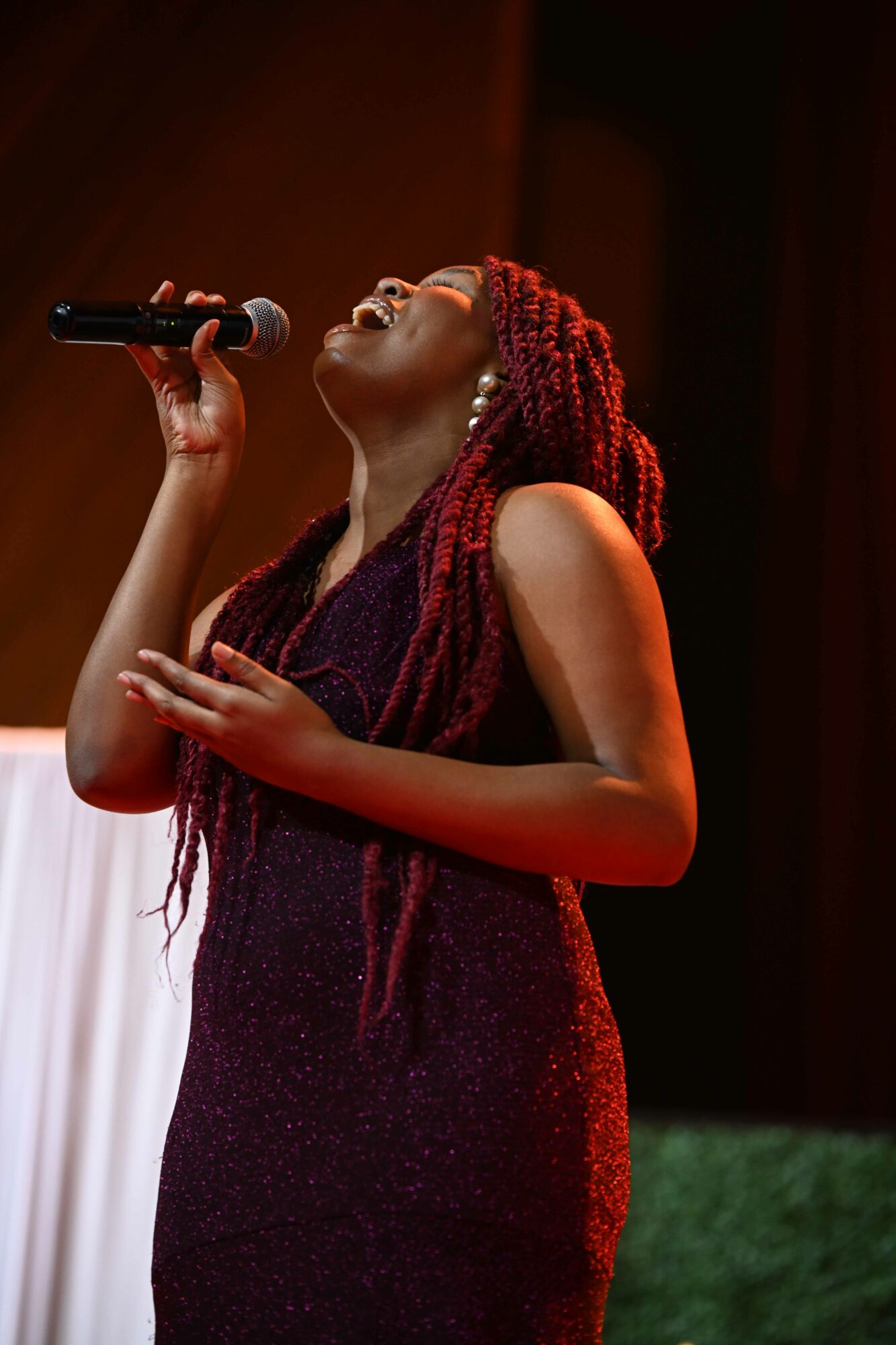 Woman singing into a microphone with red braided hair, wearing a dark dress, on stage with warm lighting.