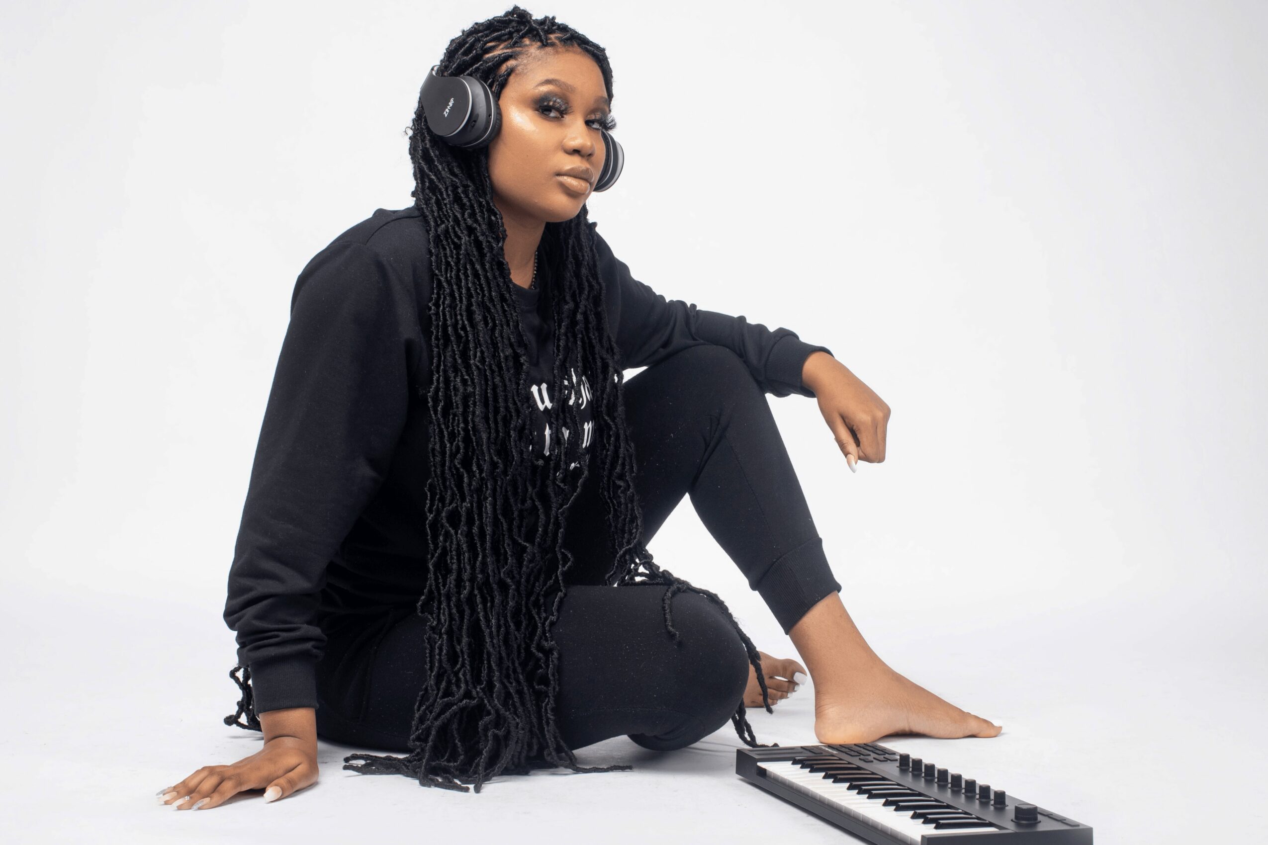 Woman with long dreadlocks wearing headphones, sitting on the floor with a keyboard, against a plain white background.