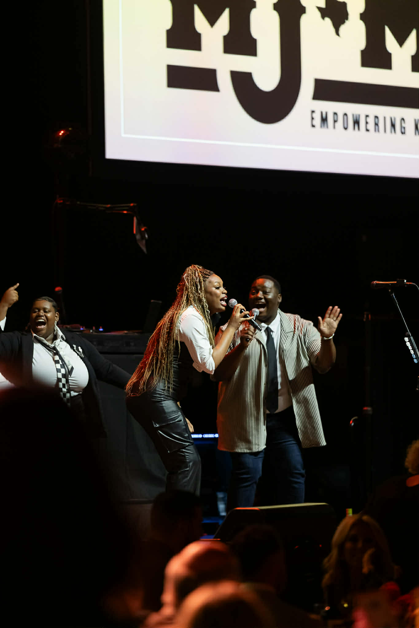 Group of people singing on stage with a large screen displaying a logo and text behind them.