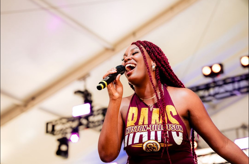 Woman singing into microphone, smiling, with stage lights and equipment in background.