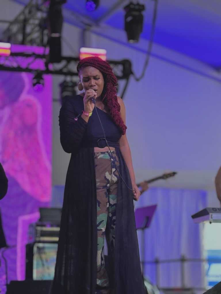 Woman singing on stage with purple and blue lighting, wearing camouflage pants and a long black coat.