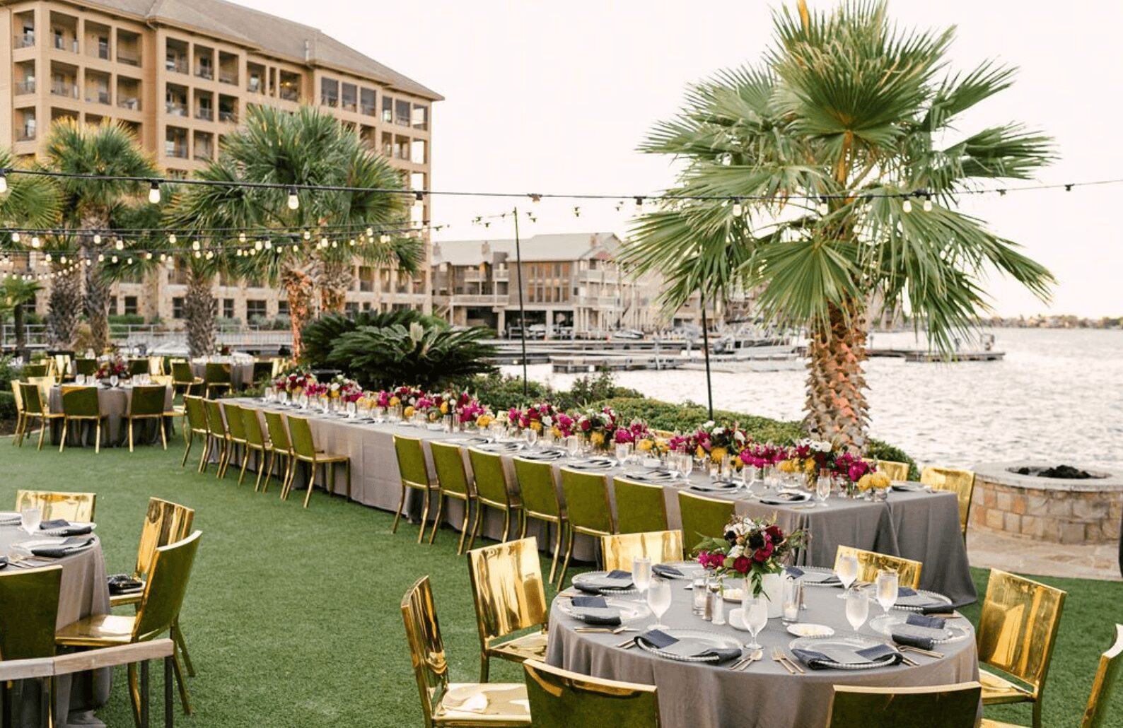 Outdoor event setup with round tables, chairs, and a long table decorated with flowers near water, palm trees, and buildings.