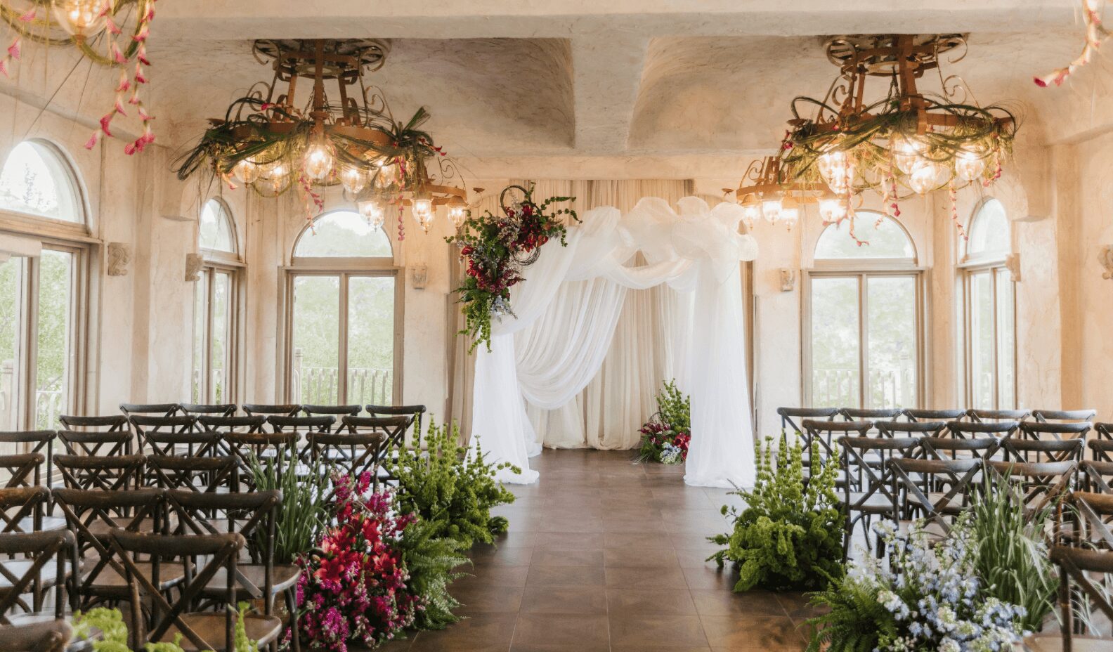 Wedding ceremony setup with chairs, floral arrangements, and a draped arch in a bright room with large windows.