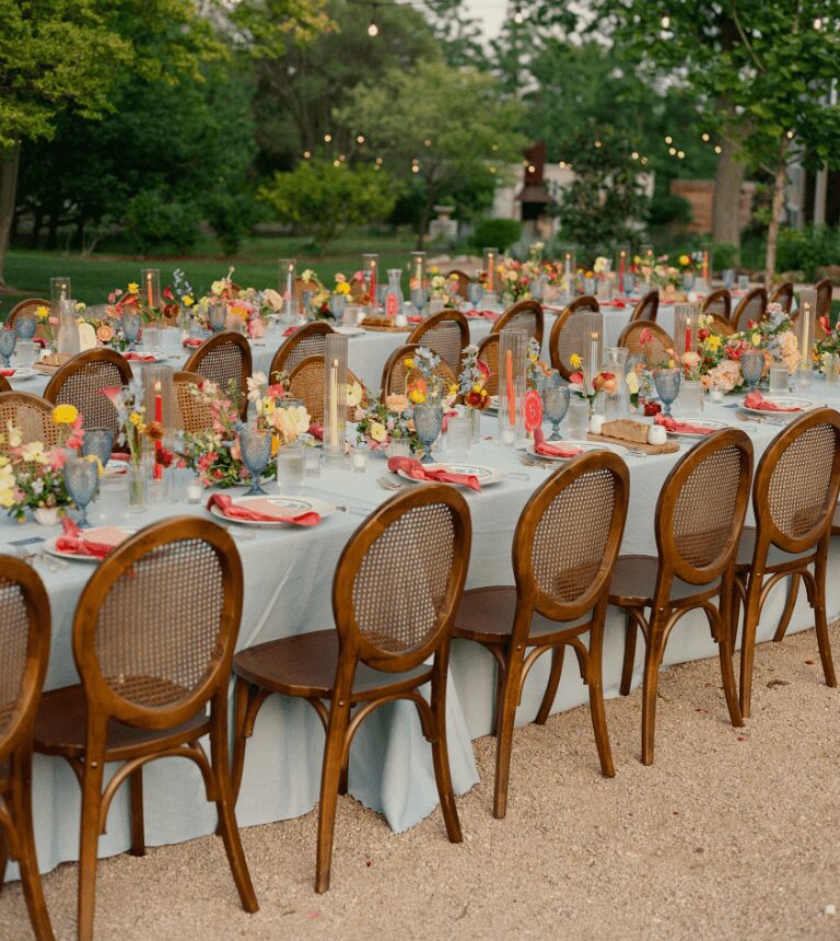 Long outdoor table set with chairs, decorated for a celebration, surrounded by trees and greenery.