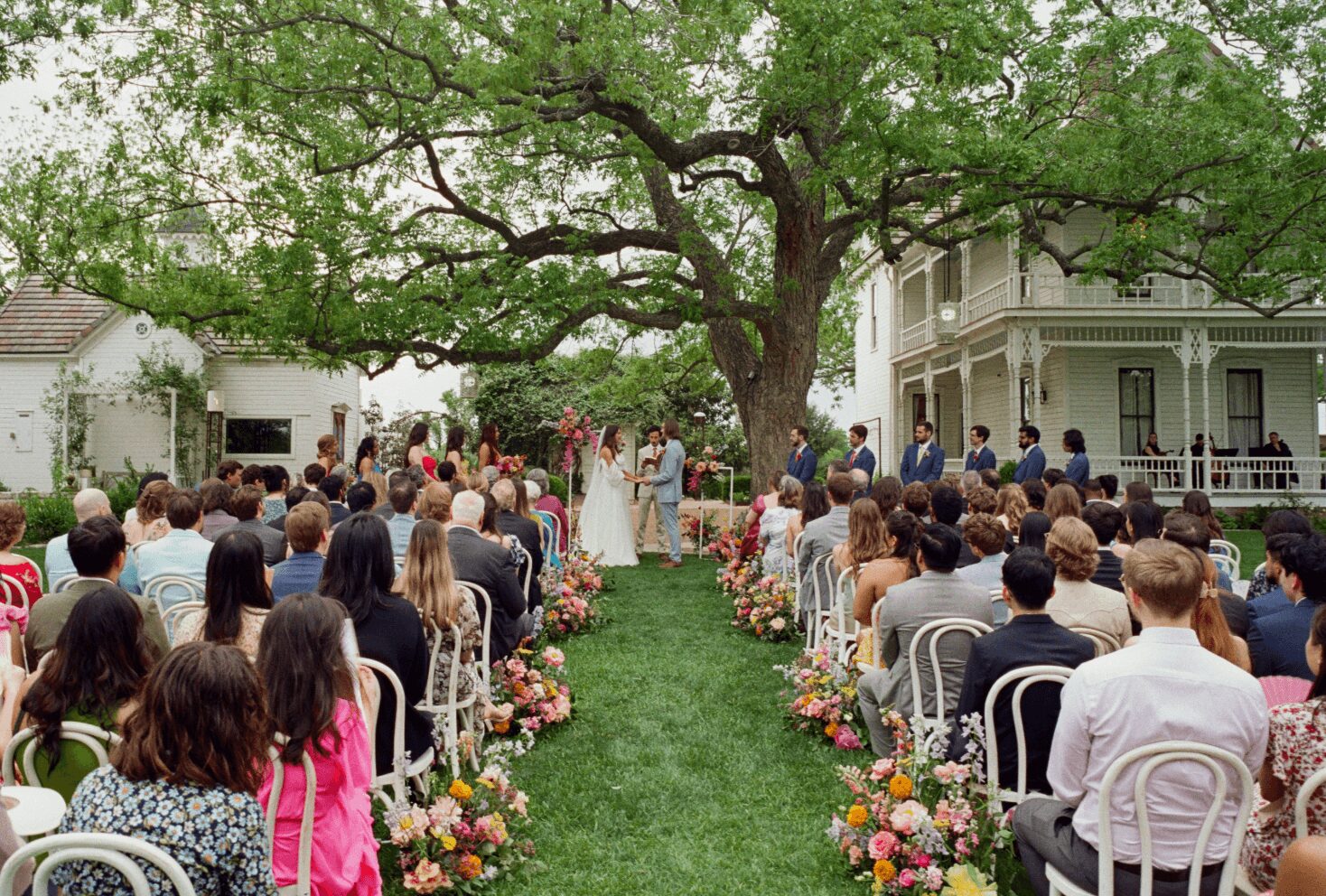 Outdoor wedding ceremony under a large tree with guests seated on both sides, decorated with flowers, in front of a white house.