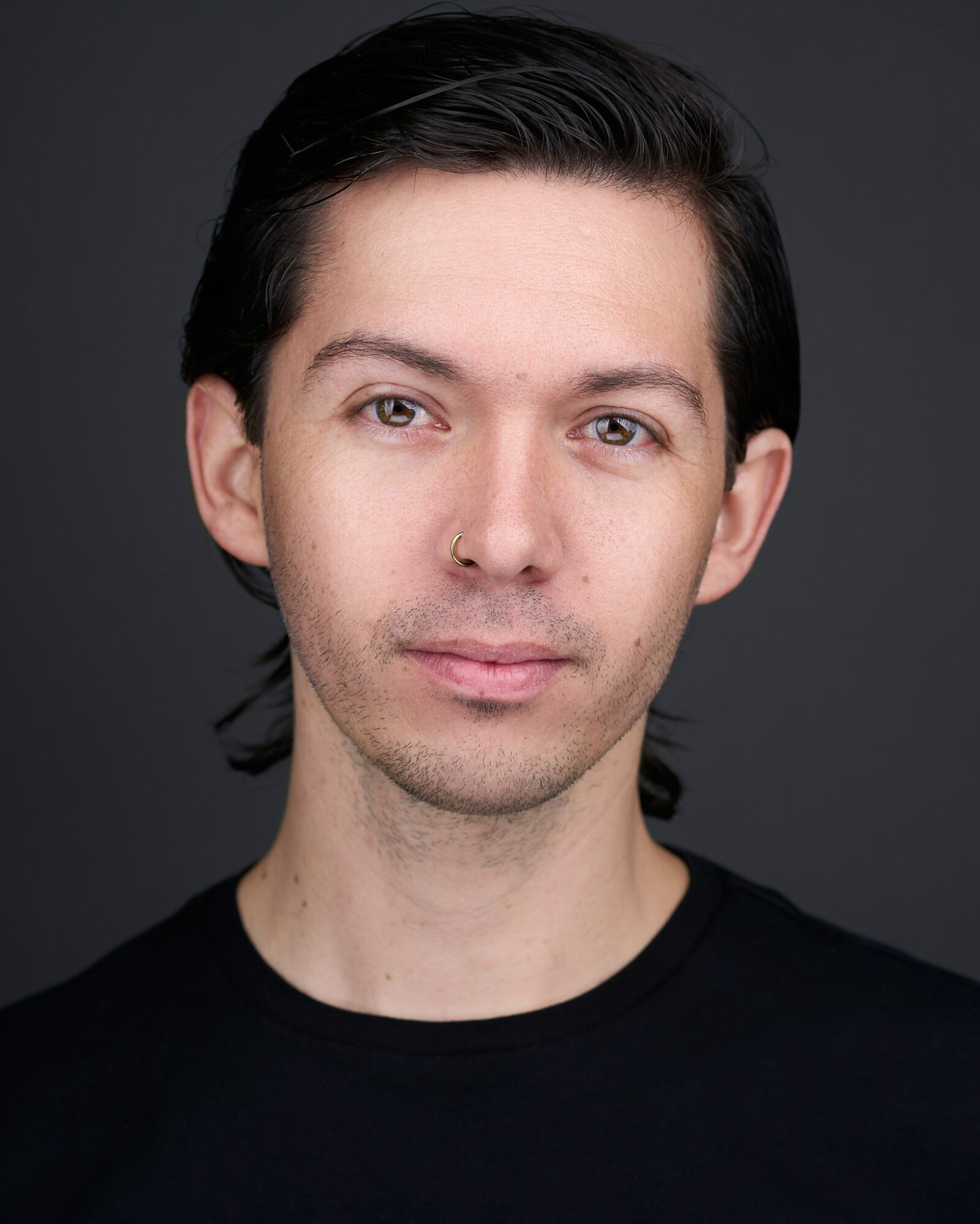 Portrait of a young man with dark hair, light skin, and a neutral expression, wearing a black shirt against a dark background.