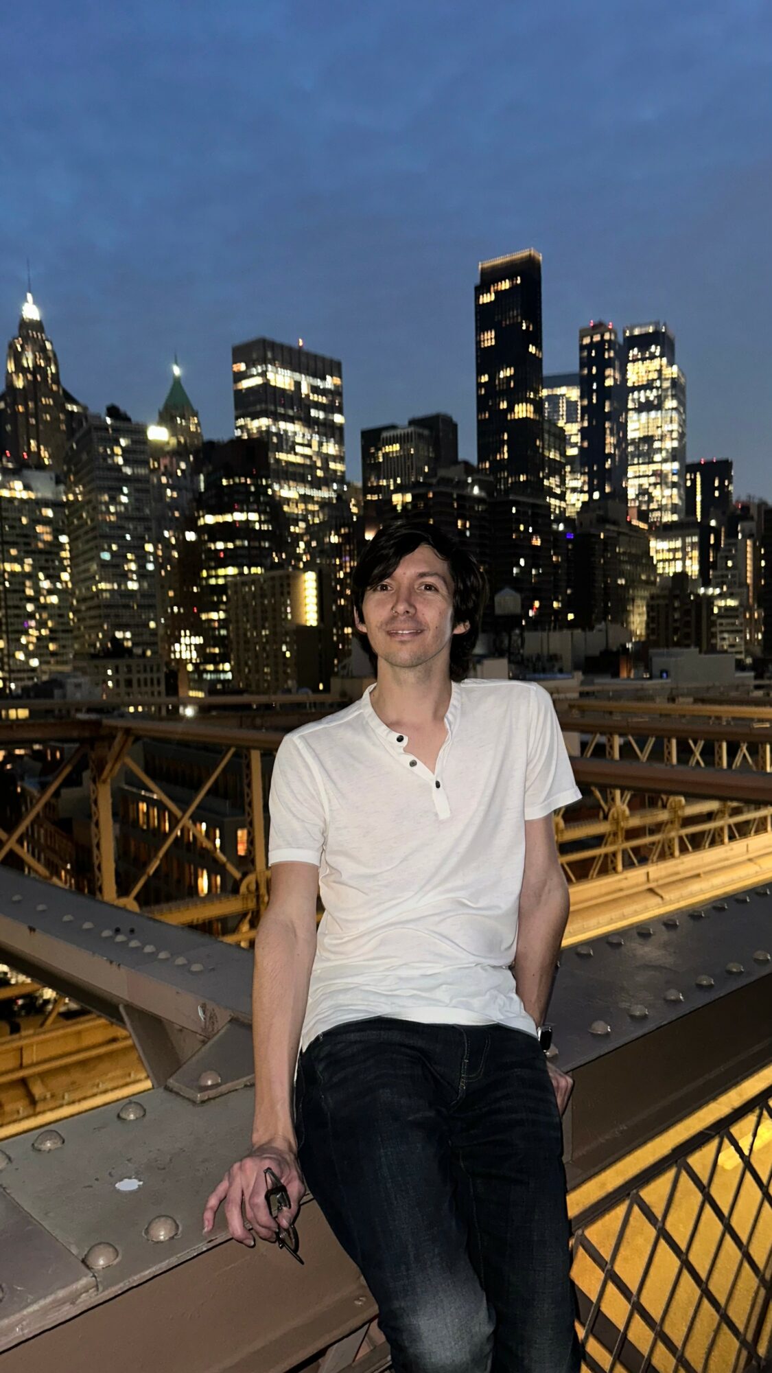 Person standing on a bridge at night with a city skyline and tall buildings illuminated in the background.
