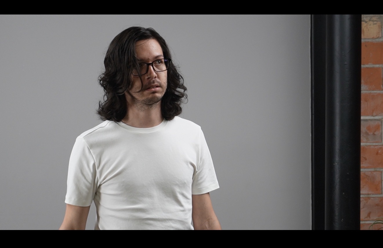 Man with long dark hair and glasses wearing a white t-shirt, standing indoors against a gray background.