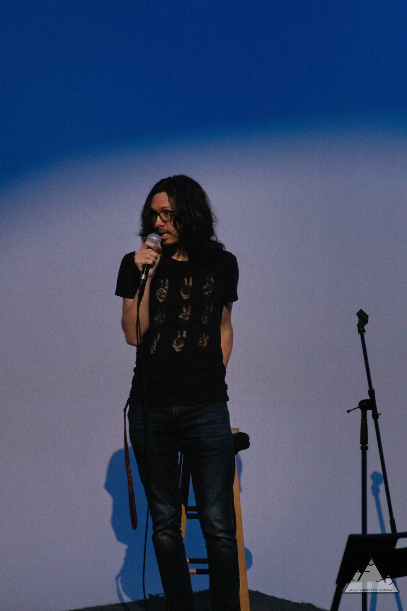 Person with curly hair and glasses speaking into a microphone on stage, with a microphone stand and chair nearby.