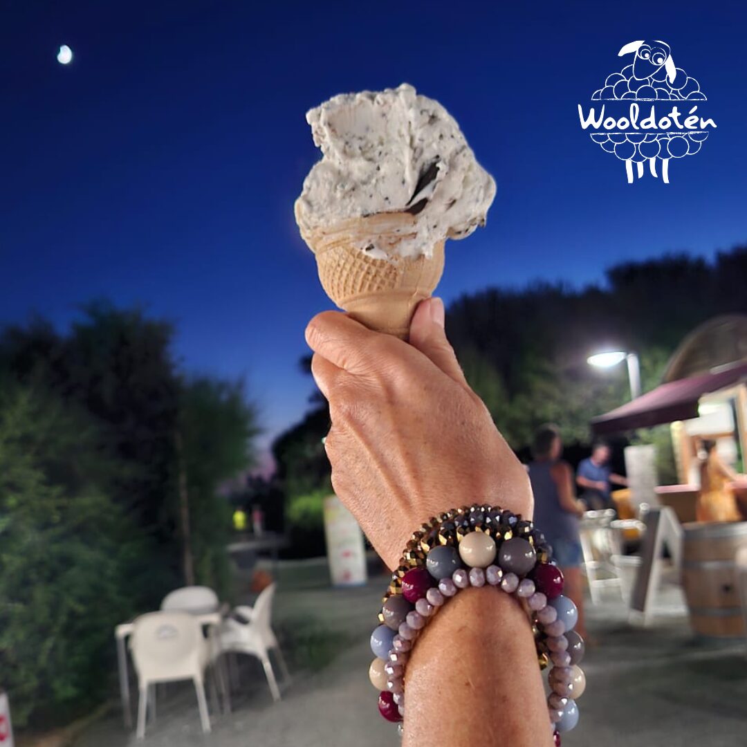 Hand holding an ice cream cone outdoors at dusk, with trees, chairs, and a building in the background.