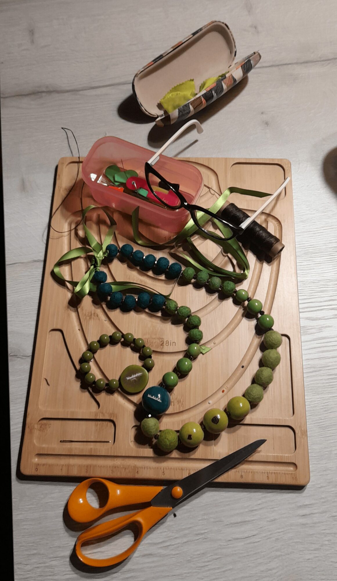 Necklaces and glasses on a wooden tray with scissors nearby, and a small open case above on a light surface.