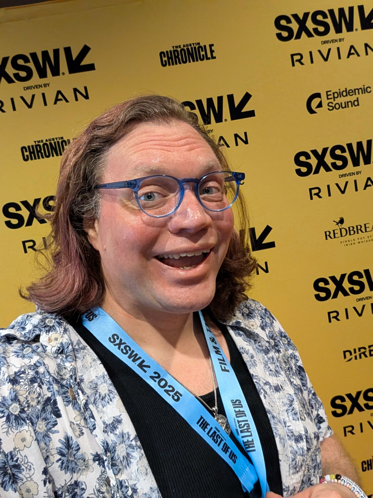 Person with long hair, glasses, smiling, wearing a patterned shirt and a blue lanyard, standing in front of a yellow SXSW backdrop.
