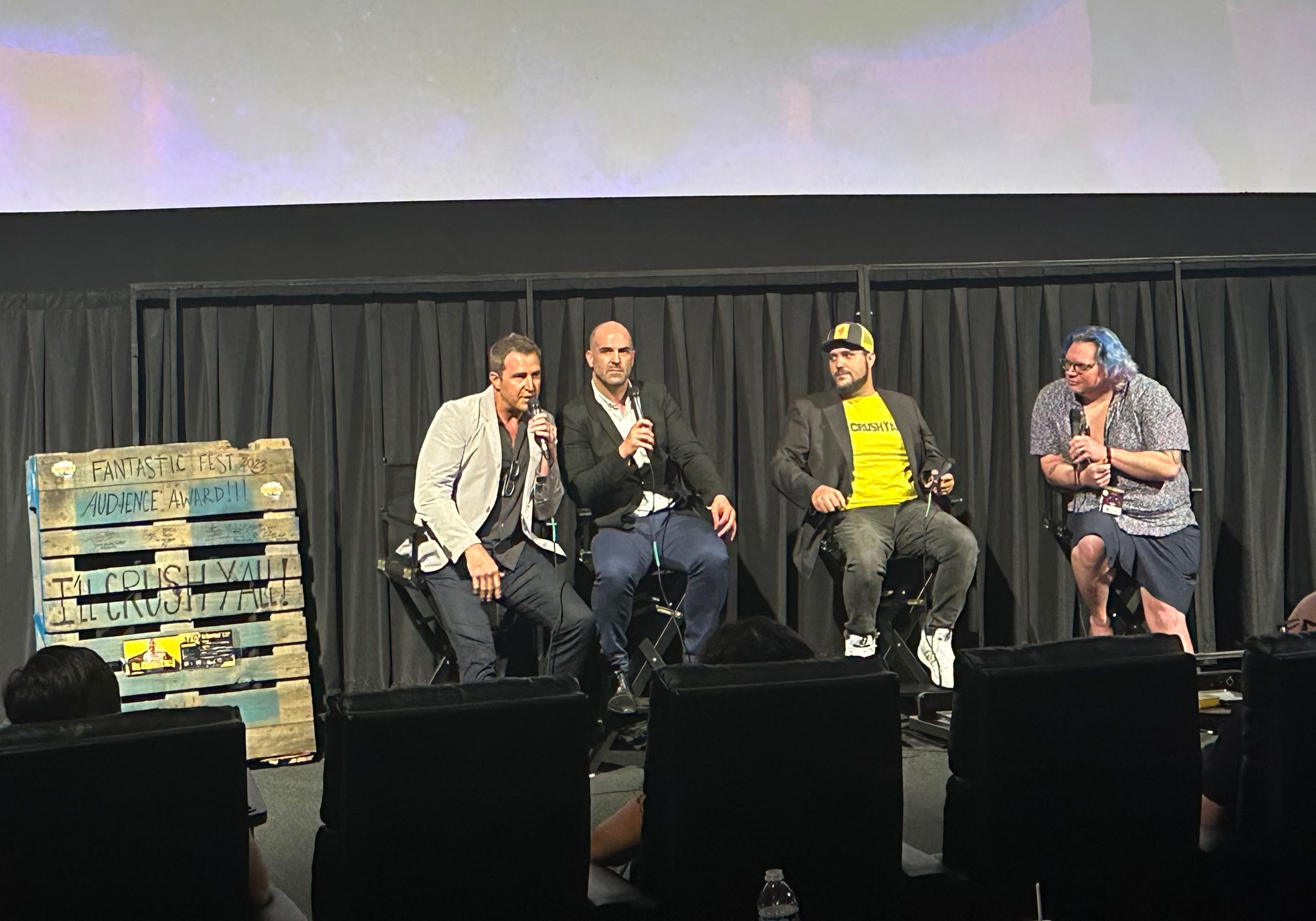 Four people sit on a stage with a black curtain background, engaged in conversation, with a wooden pallet on the left.
