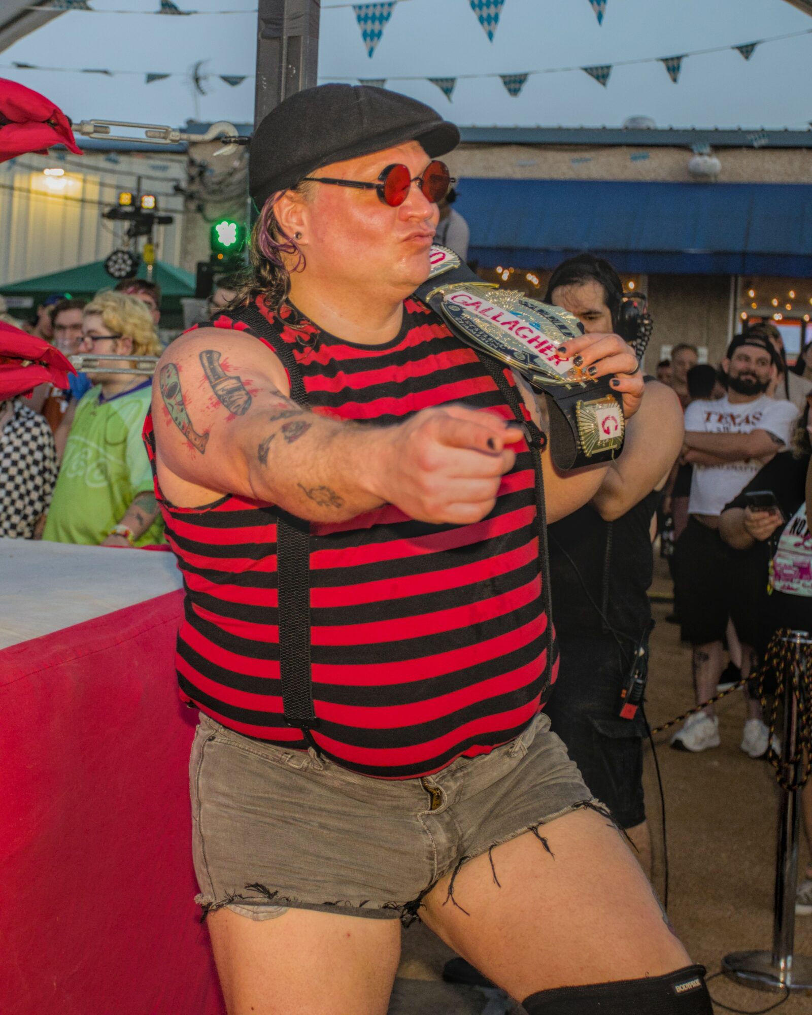 Person with tattoos, red sunglasses, black hat, striped shirt, and shorts holding a bottle at an outdoor event.