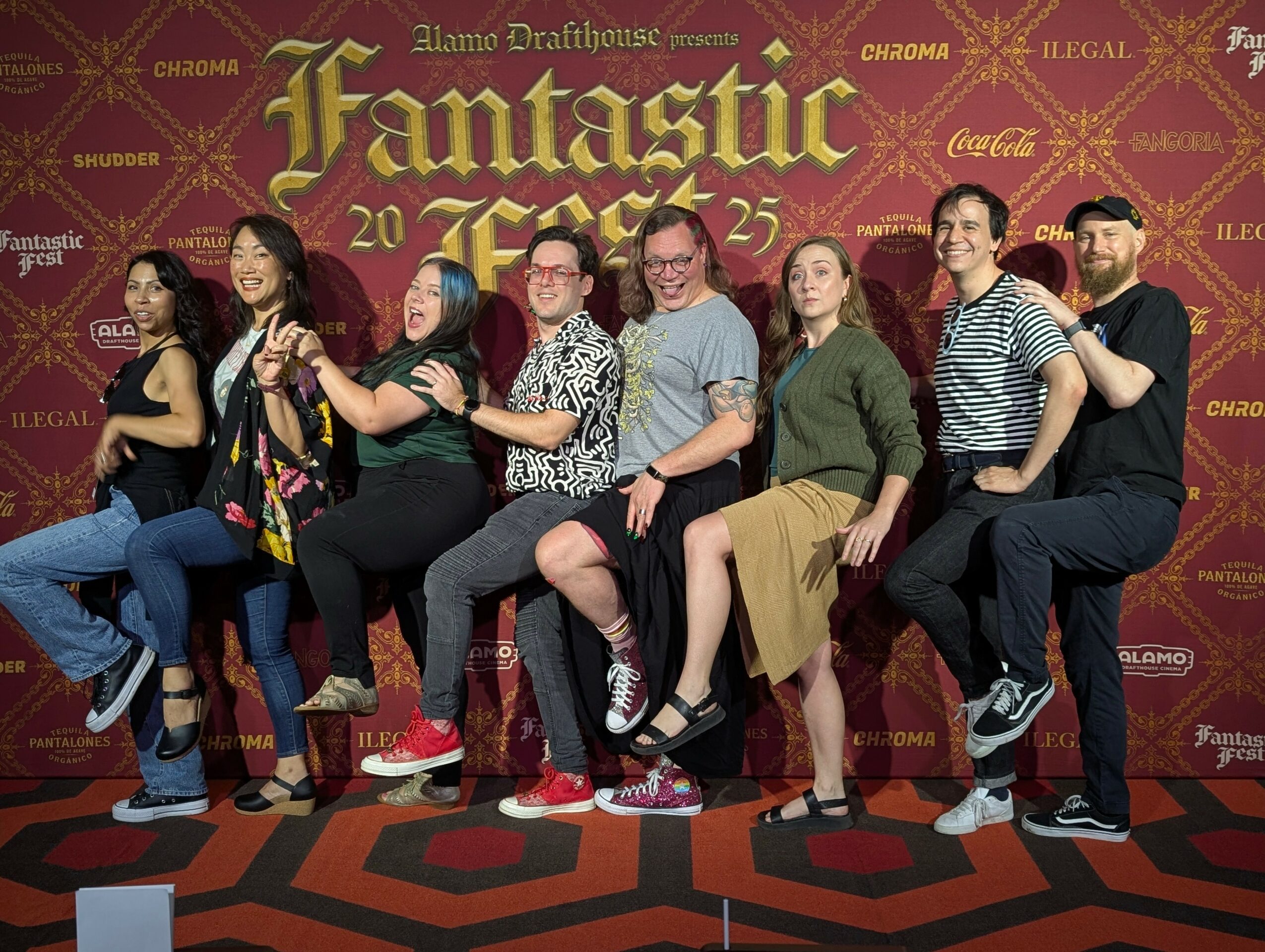 Group of nine people sitting on a bench in front of a red backdrop with yellow text, smiling and posing together.
