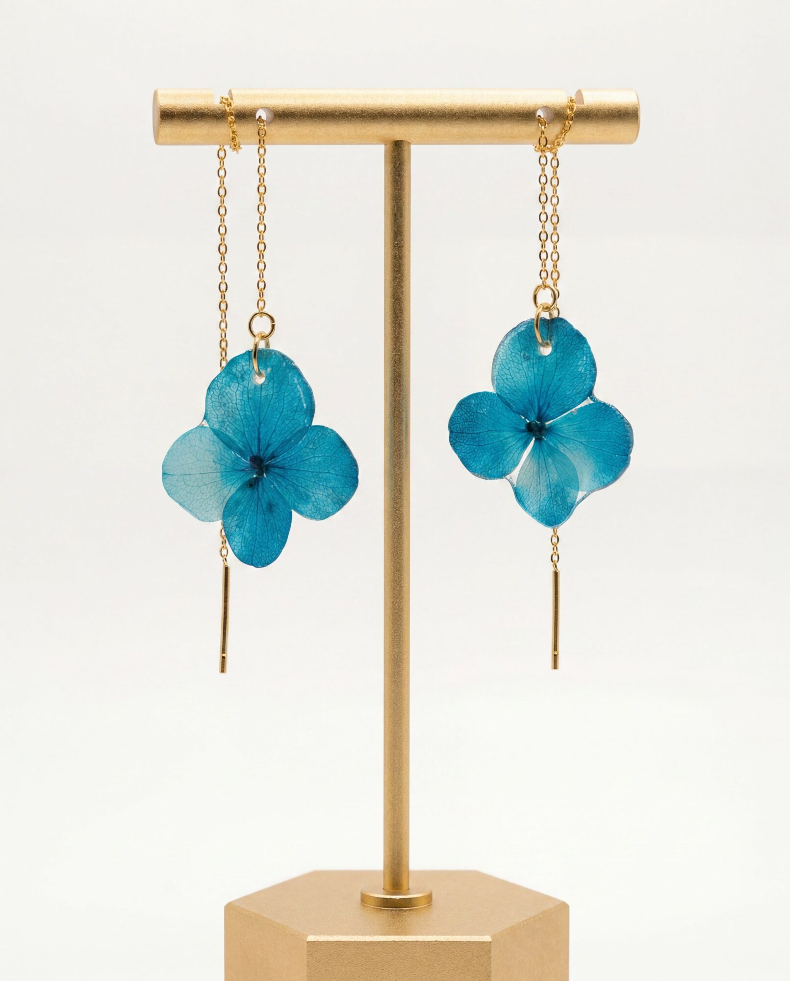 Blue flower-shaped earrings hanging from a gold display stand, with a white background.