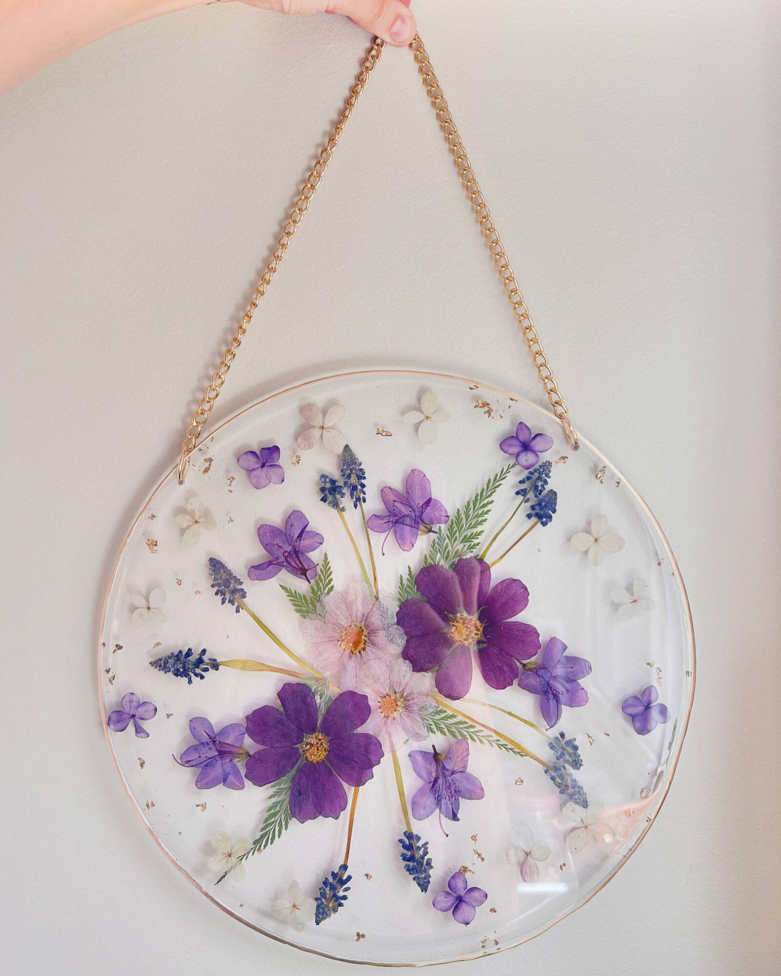 Round bag with purple and pink flowers and green leaves, hanging by a gold chain, against a plain background.